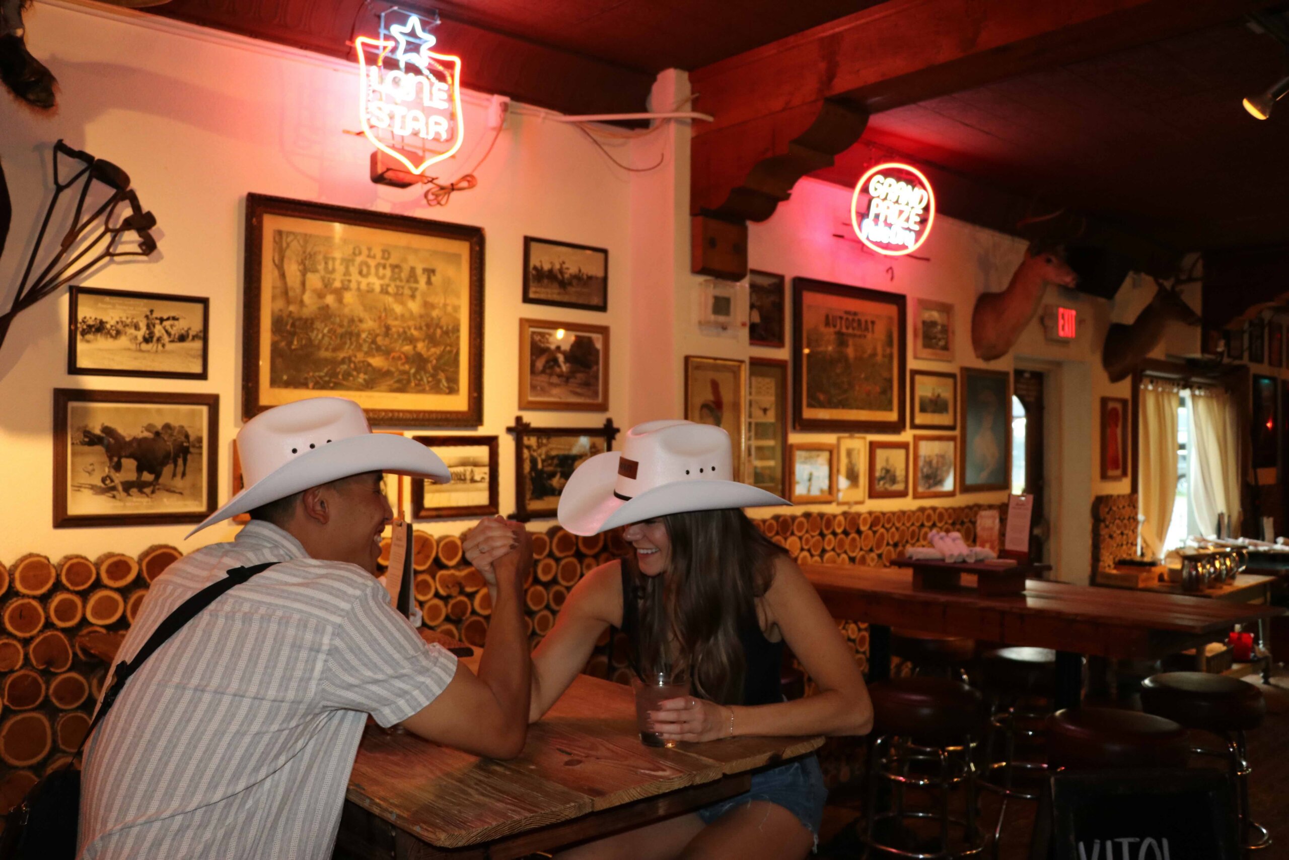 CRAFT Beer Market Two people in white cowboy hats arm wrestle at a wooden table inside a western-themed bar decorated with neon signs and framed photos.