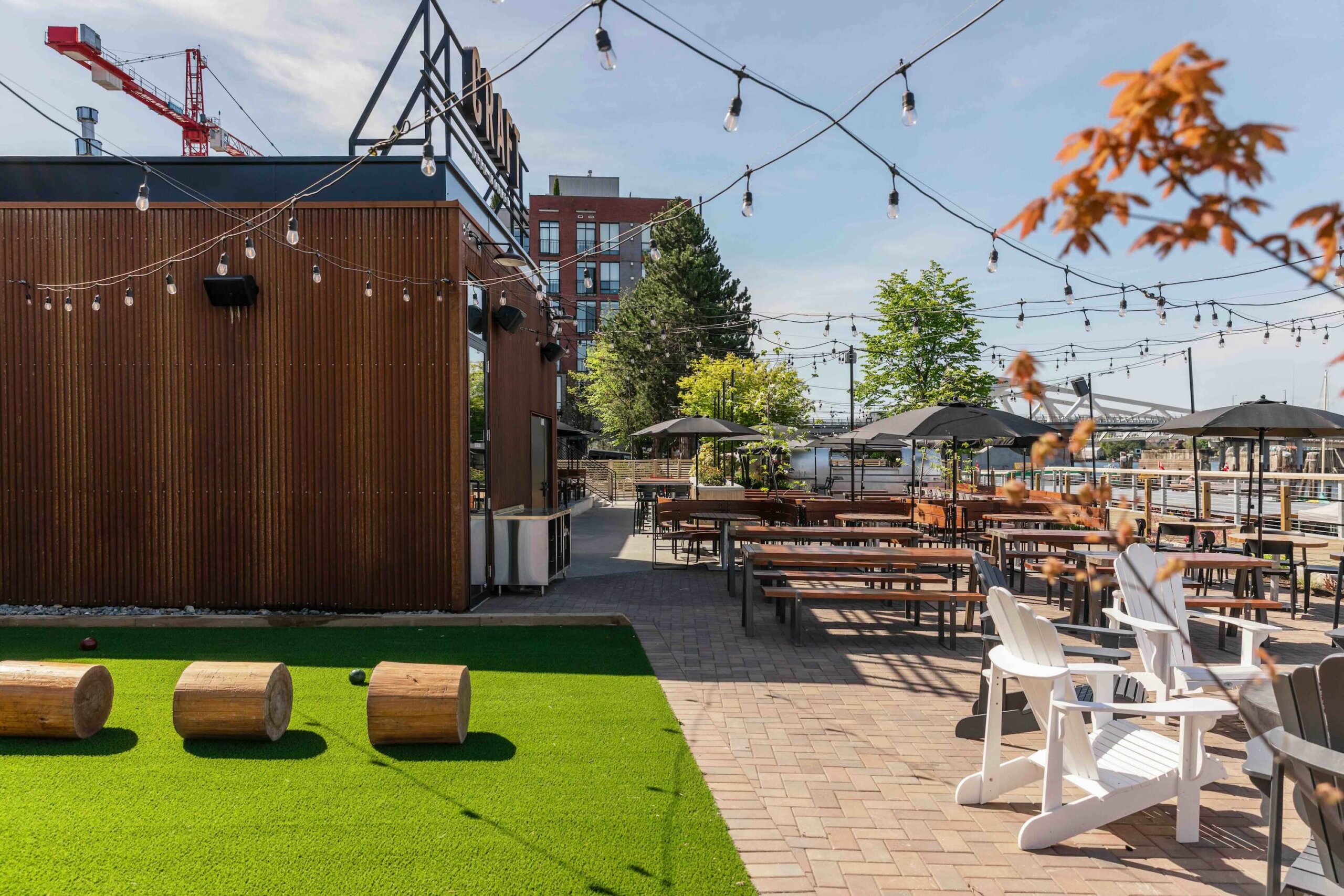 CRAFT Beer Market Outdoor patio area with picnic tables, string lights, artificial turf, wooden logs, and white chairs; enjoy craft cocktails amid social vibes with umbrellas and trees in the background under a clear sky.