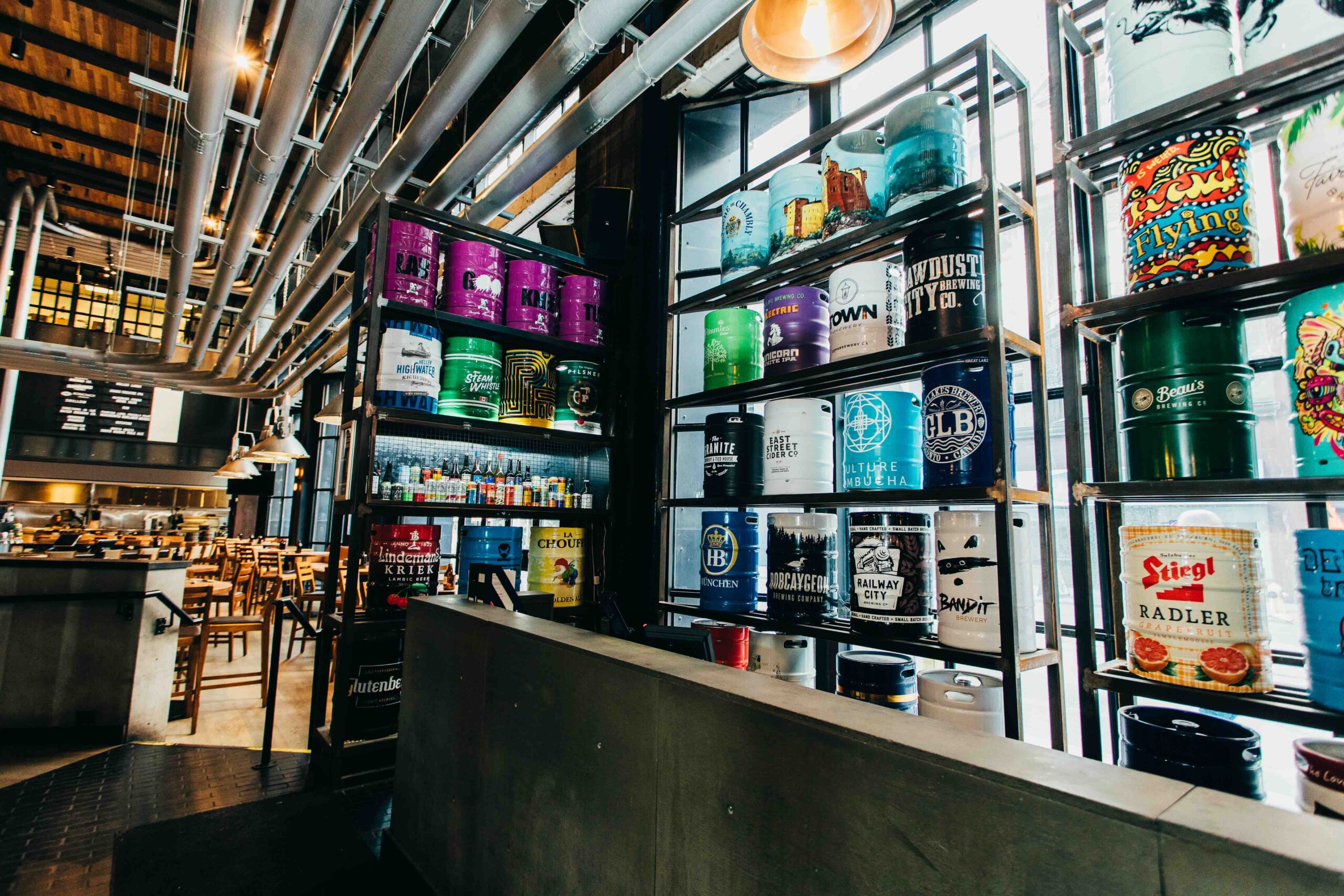 CRAFT Beer Market Interior of a modern bar with shelves displaying colorful beer kegs and cans, industrial pipes on the ceiling, and wooden tables and chairs in the background.