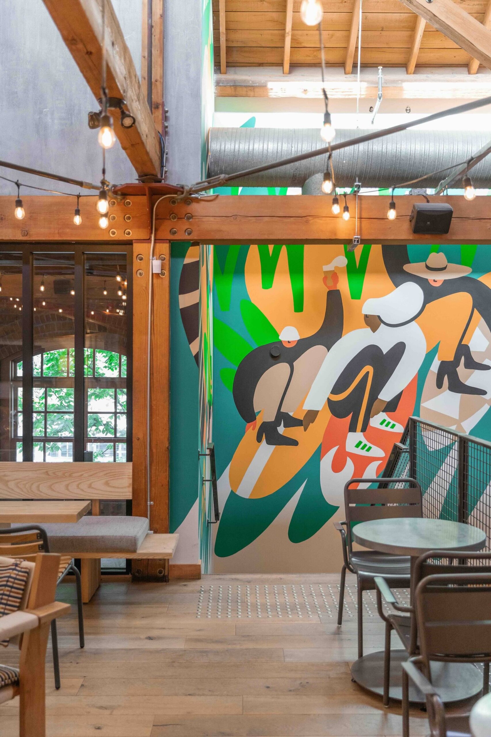 CRAFT Beer Market A modern café interior with wooden beams, string lights, and a colorful mural depicting abstract cyclists on the wall. Enjoy craft cocktails while soaking in the social vibes among tables and chairs arranged throughout the space.