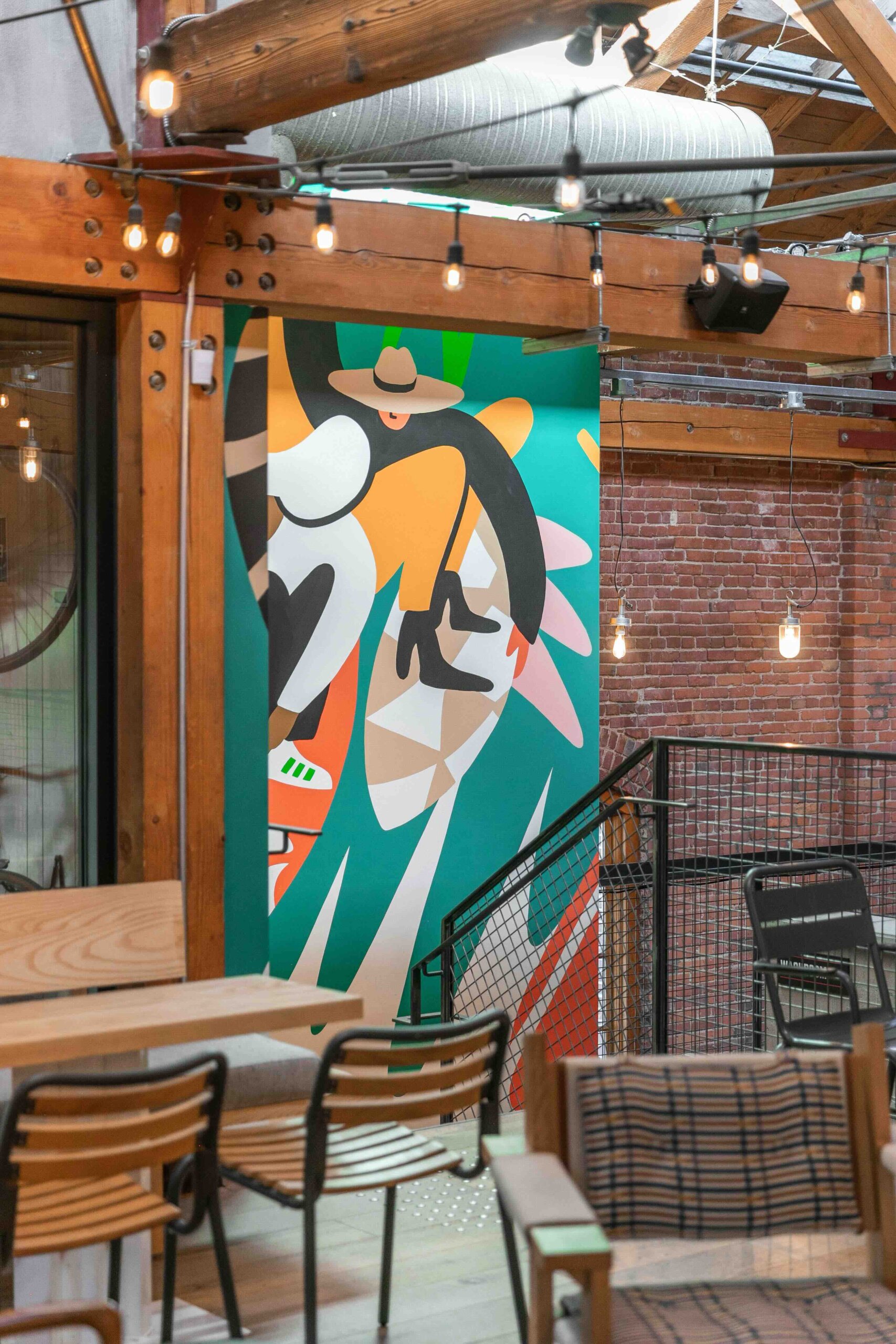 CRAFT Beer Market Outdoor beer garden with wooden chairs and tables, string lights, and a colorful mural featuring abstract shapes and a person in a hat on a brick wall—perfect for enjoying craft beer or cocktails.