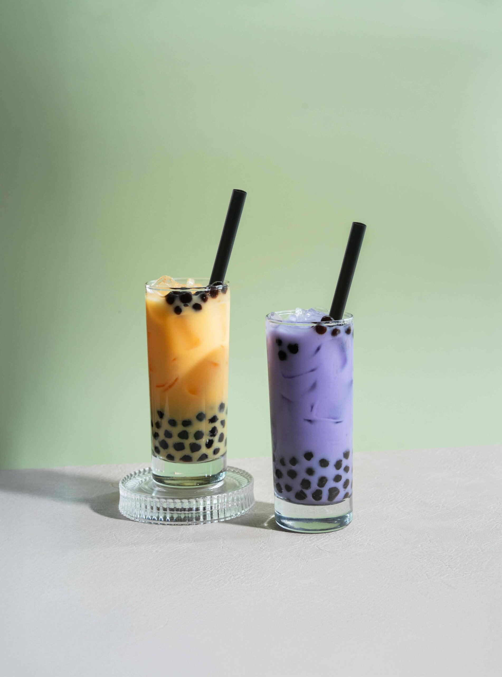 CRAFT Beer Market Two tall glasses of bubble tea with black straws, one orange and one purple, filled with black tapioca pearls, on a light surface against a pale green background—perfect for sipping alongside craft beer in a lively beer garden.