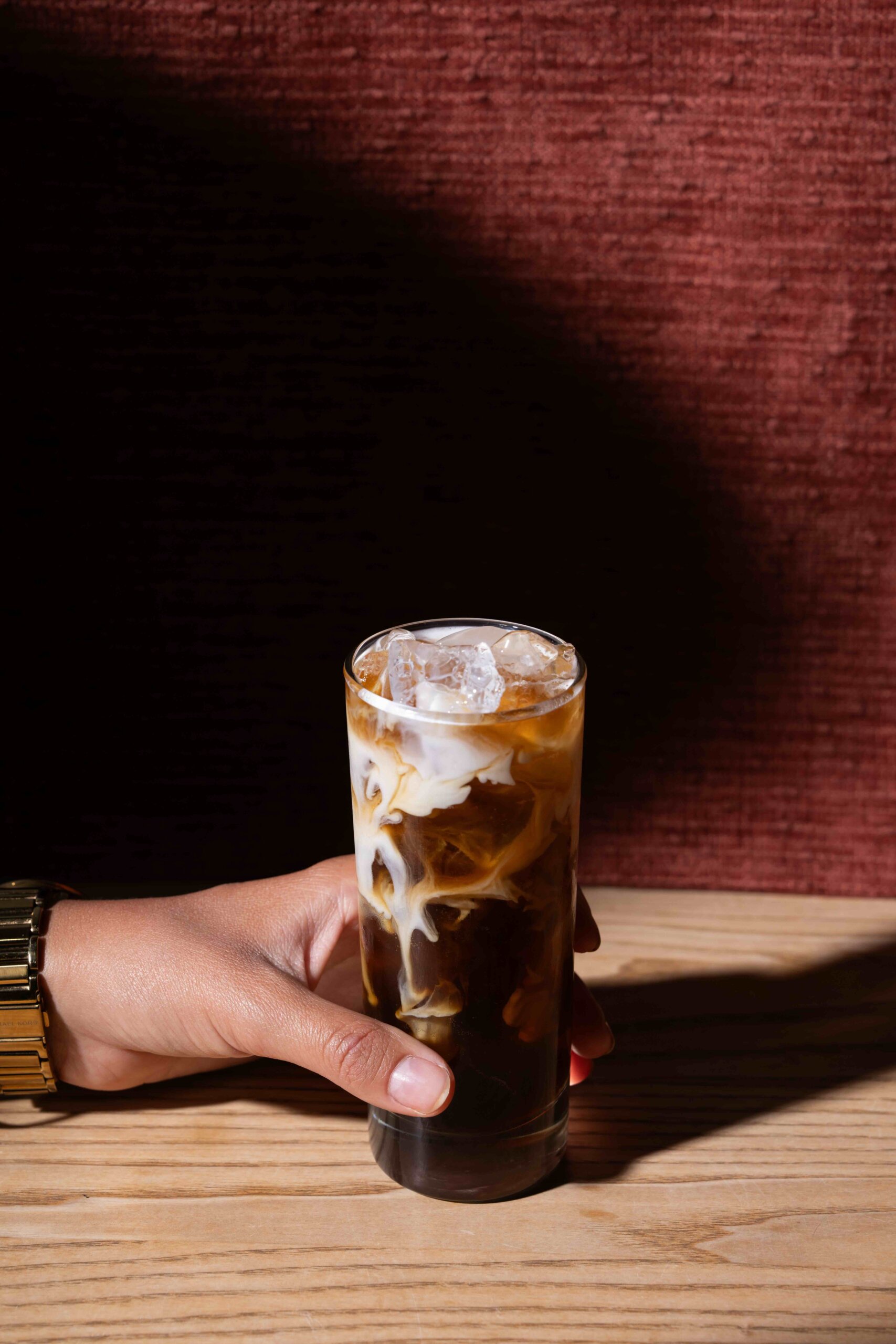 CRAFT Beer Market A hand holding a glass of iced coffee with milk swirling through it, on a wooden table against a red textured background.