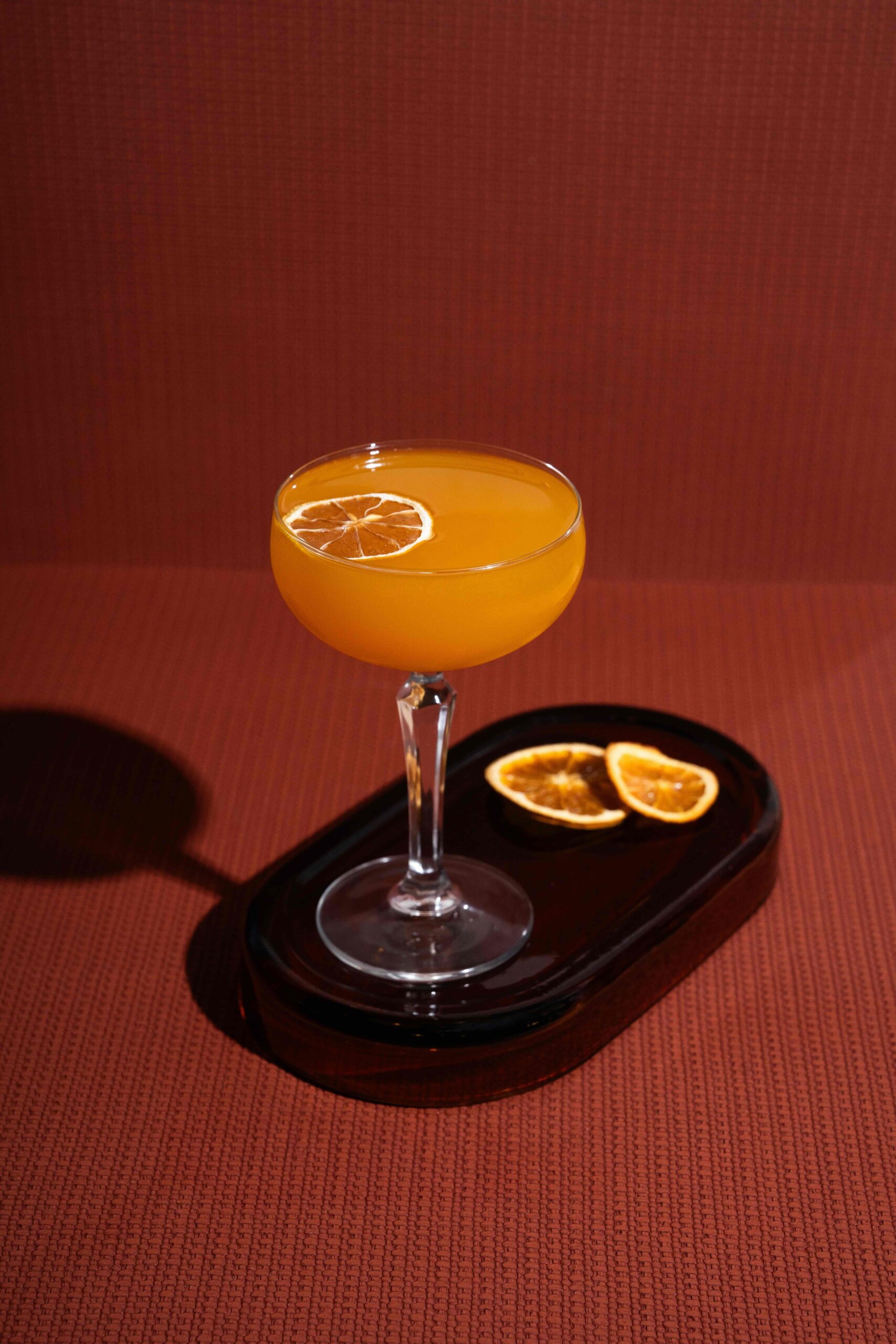 CRAFT Beer Market A cocktail in a coupe glass garnished with a dried orange slice, placed on a black tray with two more dried orange slices, against a textured brown background.