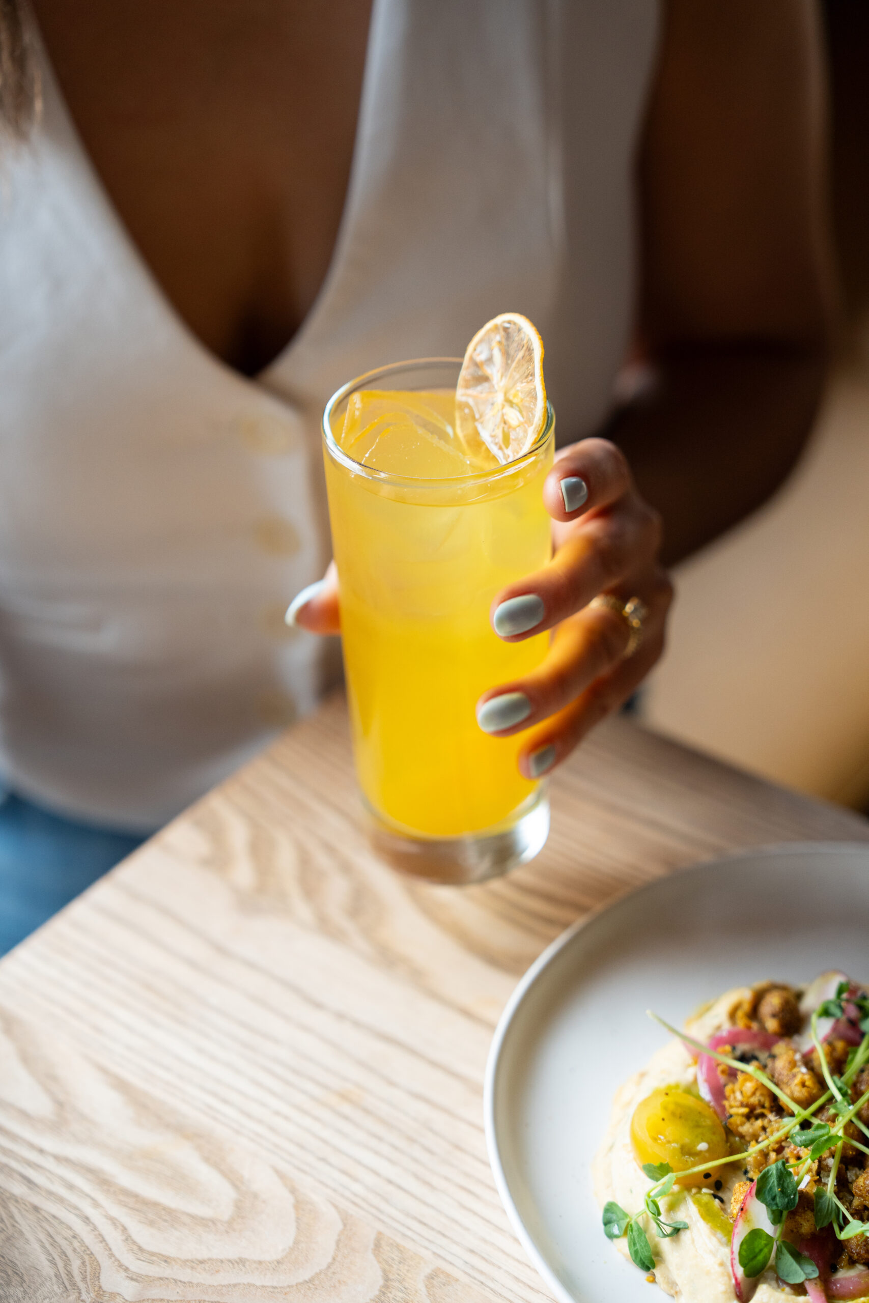 CRAFT Beer Market A person holding a glass of yellow citrus drink with a lemon slice garnish sits at a table with a plate of food.