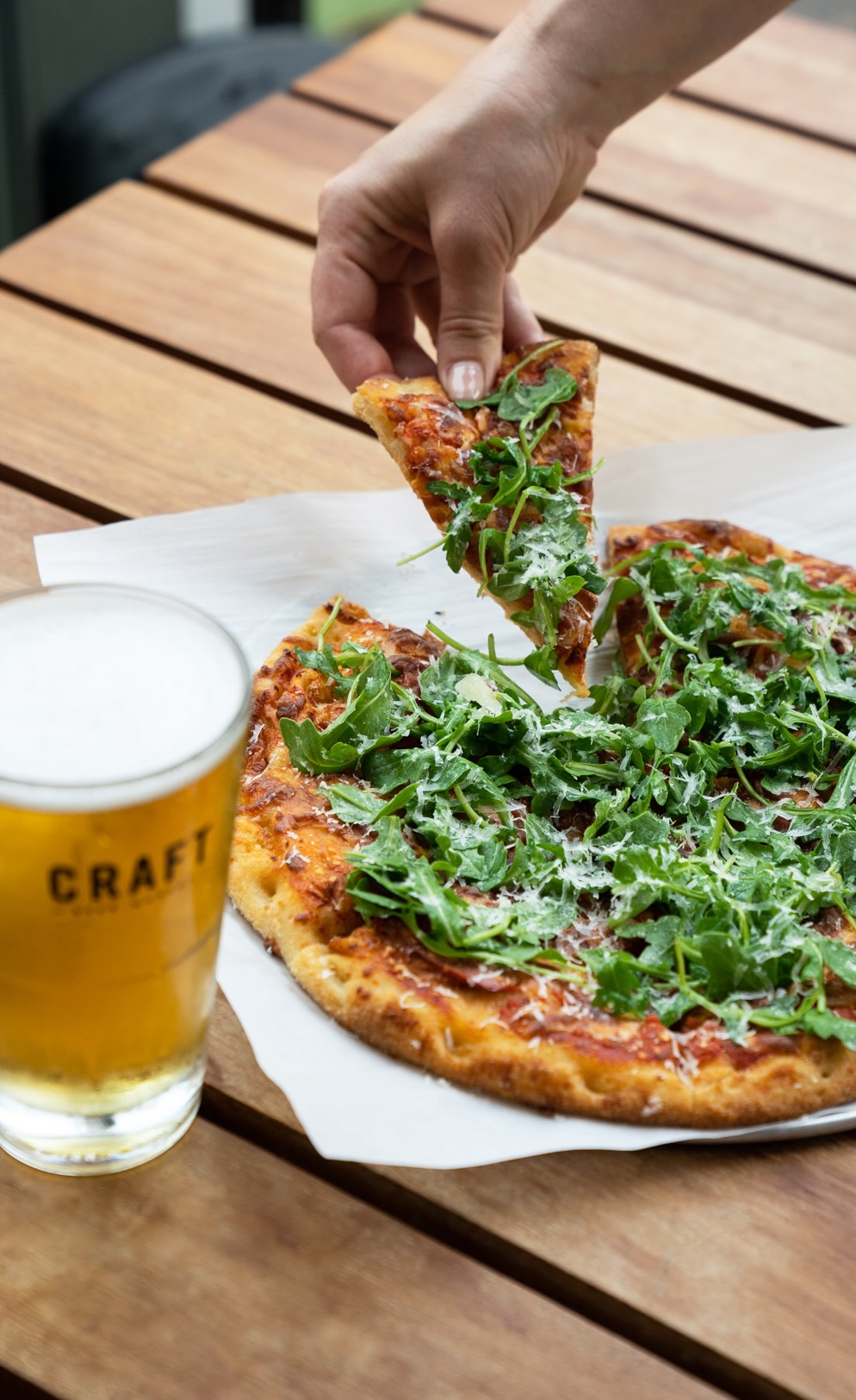 CRAFT Beer Market A hand takes a slice of pizza topped with arugula from a whole pizza on a wooden table, next to a glass of beer labeled "CRAFT"—perfect for social vibes and casual dining in a lively beer garden setting.