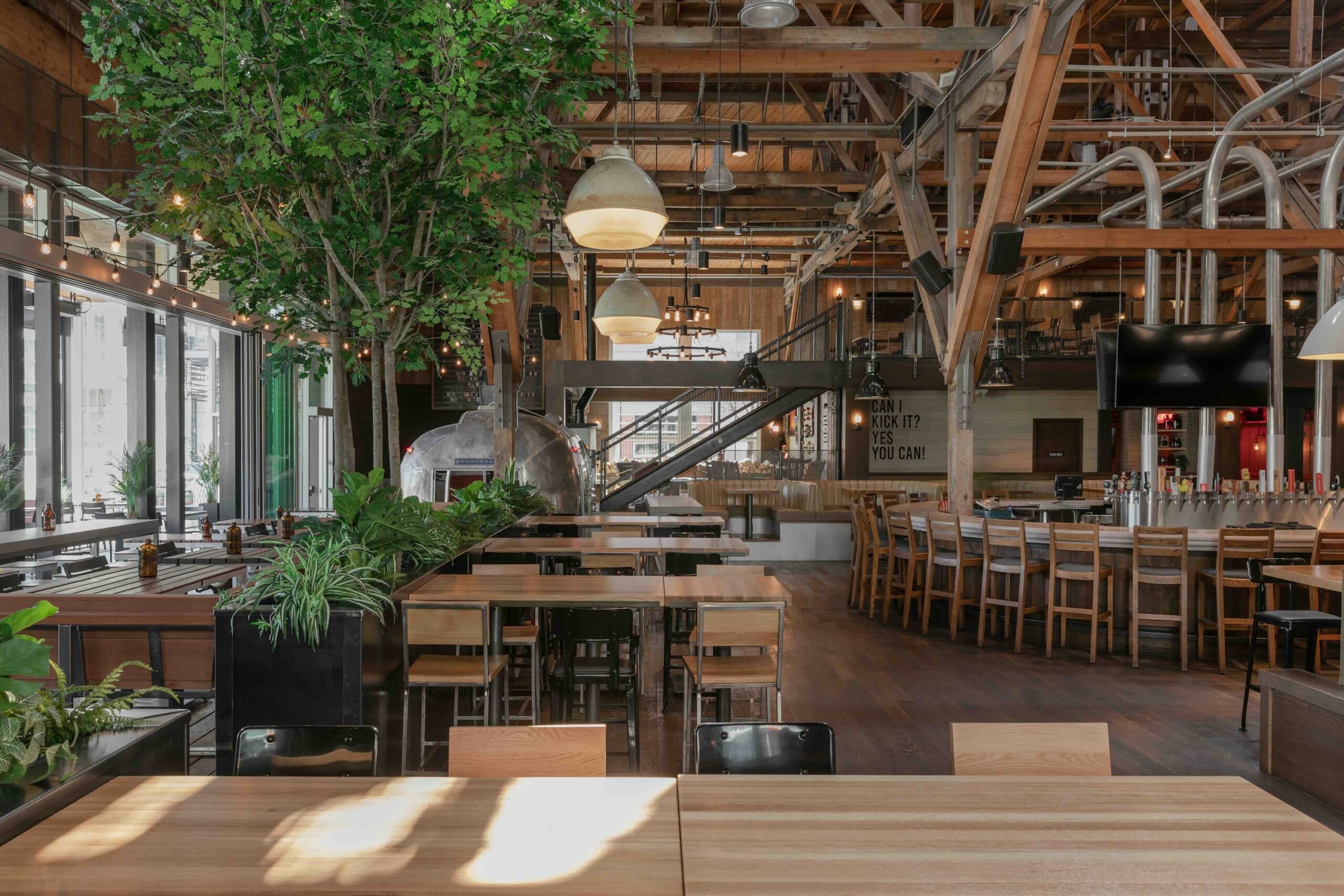 CRAFT Beer Market Spacious restaurant interior with wooden tables and chairs, a row of barstools for craft cocktails, large indoor plants, and exposed beams and pipes create inviting social vibes.