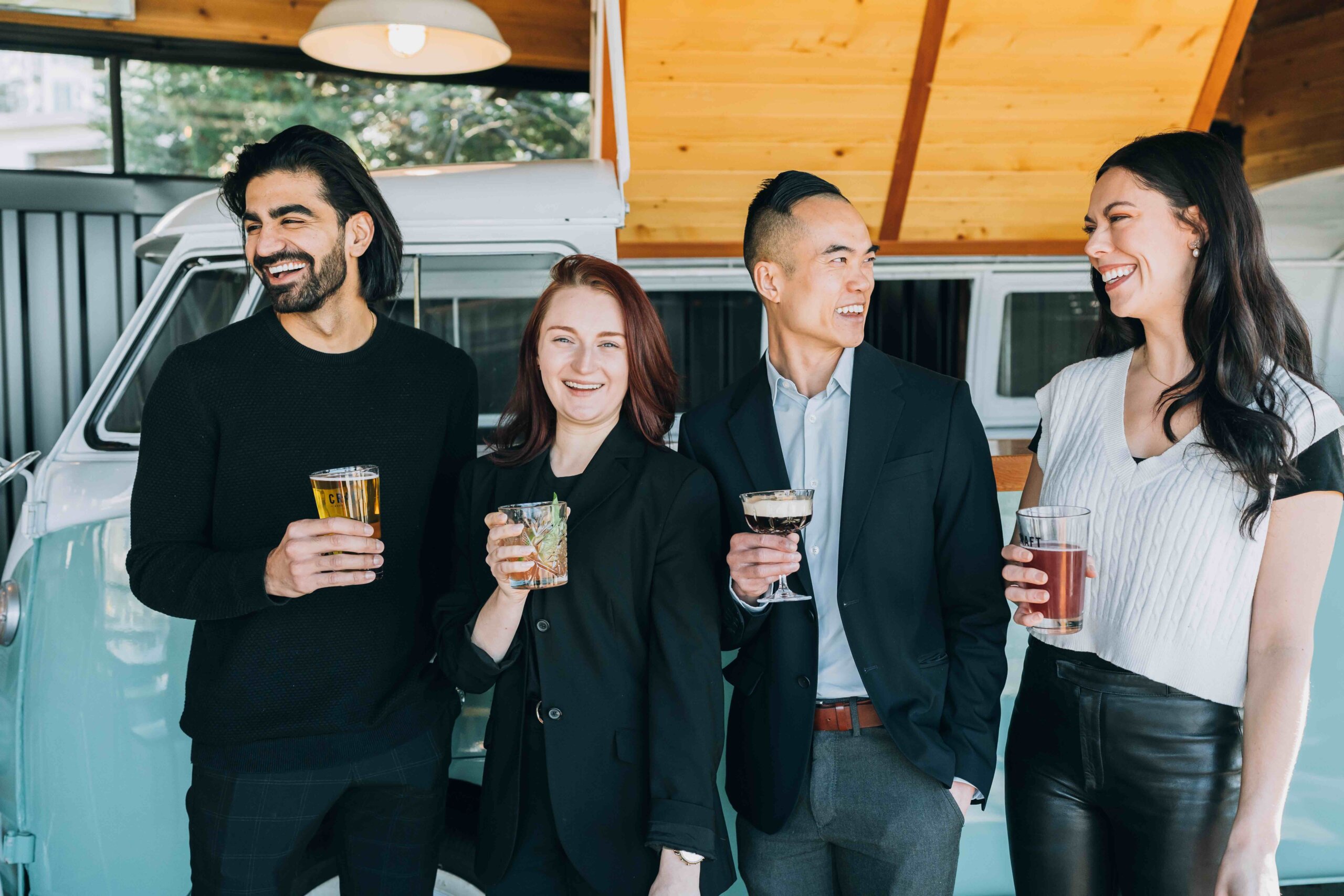 CRAFT Beer Market Four people stand in front of a retro van, smiling and holding drinks, dressed in semi-formal attire in a bright, modern setting.