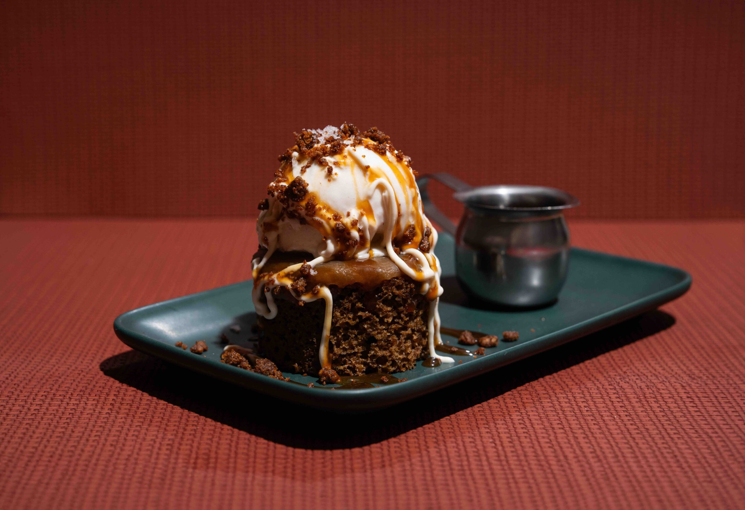 CRAFT Beer Market A dessert of ice cream and caramel sauce on a square piece of cake, topped with crumbles, is served on a black plate beside a metal sauce pitcher on a red background.