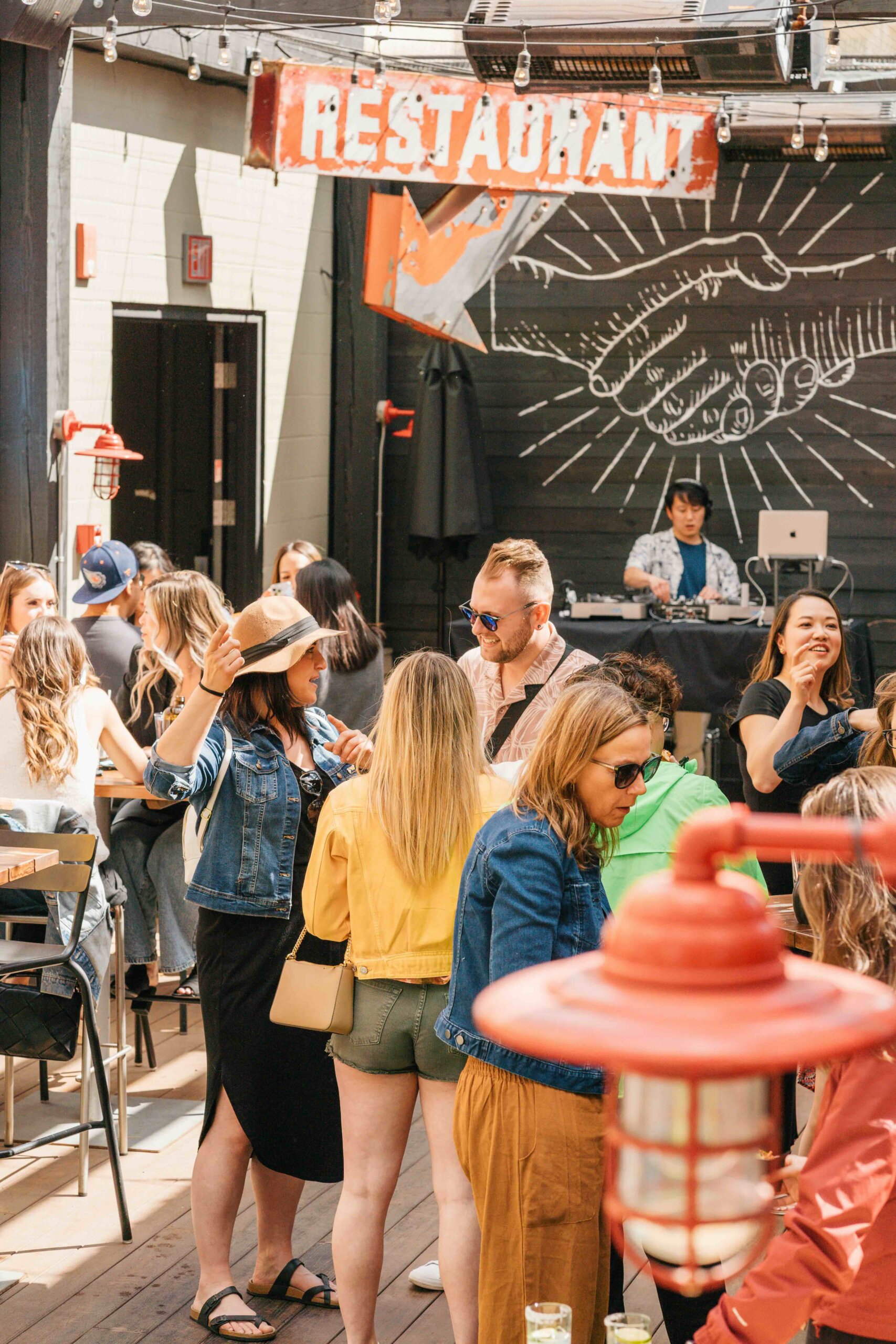 CRAFT Beer Market People socializing and standing in an outdoor restaurant with a painted mural, tables, and a red “RESTAURANT” sign overhead.