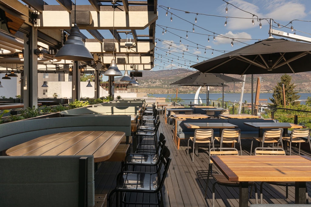 CRAFT Beer Market Outdoor restaurant patio with wooden tables, chairs, string lights, and umbrellas overlooking a lake and distant mountains under a partly cloudy sky.