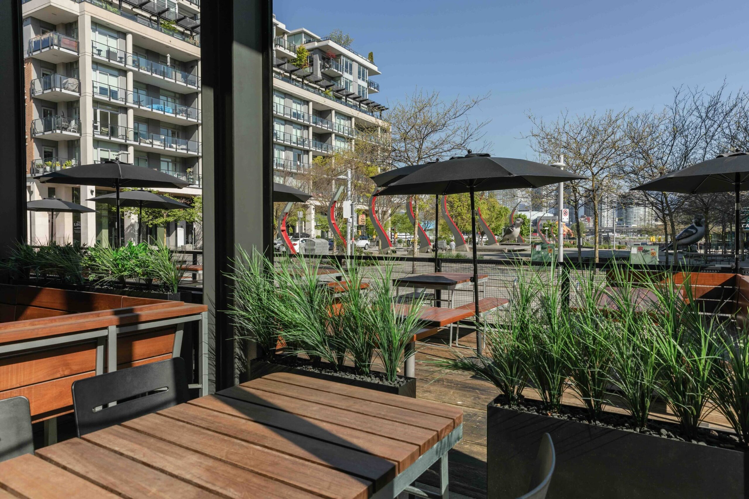 CRAFT Beer Market Outdoor patio seating with wooden tables, black chairs, planter boxes with tall grass, and black umbrellas; apartment buildings and trees visible in the background.
