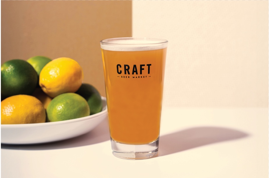 CRAFT Beer Market A glass of craft beer with a foamy head sits on a white surface next to a bowl of lemons and limes, setting the scene for perfect social vibes.