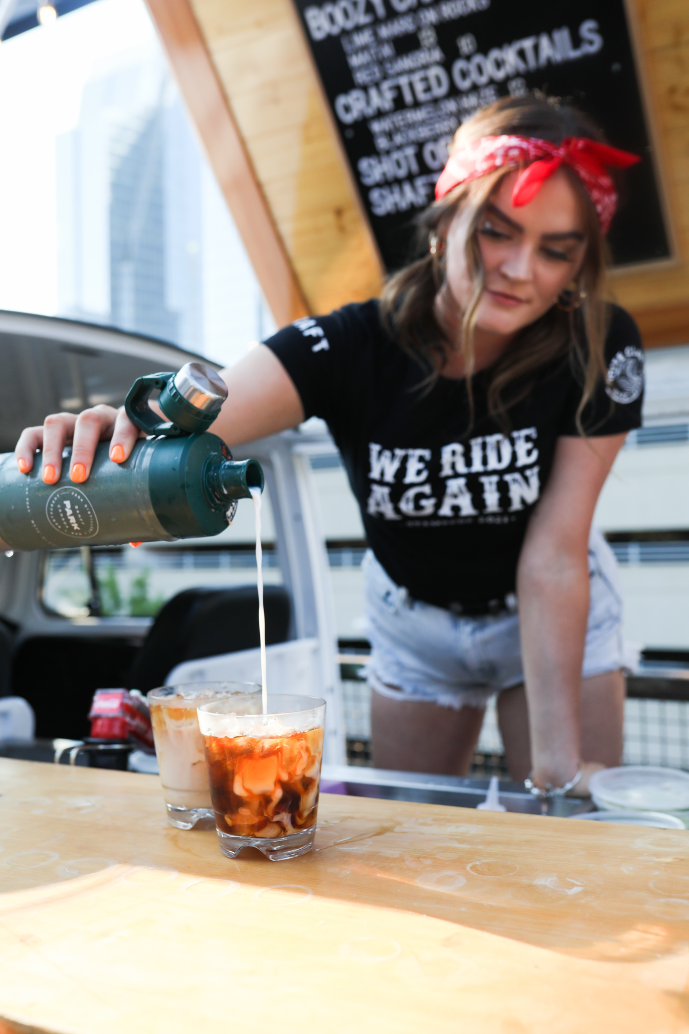 CRAFT Beer Market A woman pours cream into iced coffee drinks made with locally sourced ingredients at an outdoor stand, with a cityscape in the background.