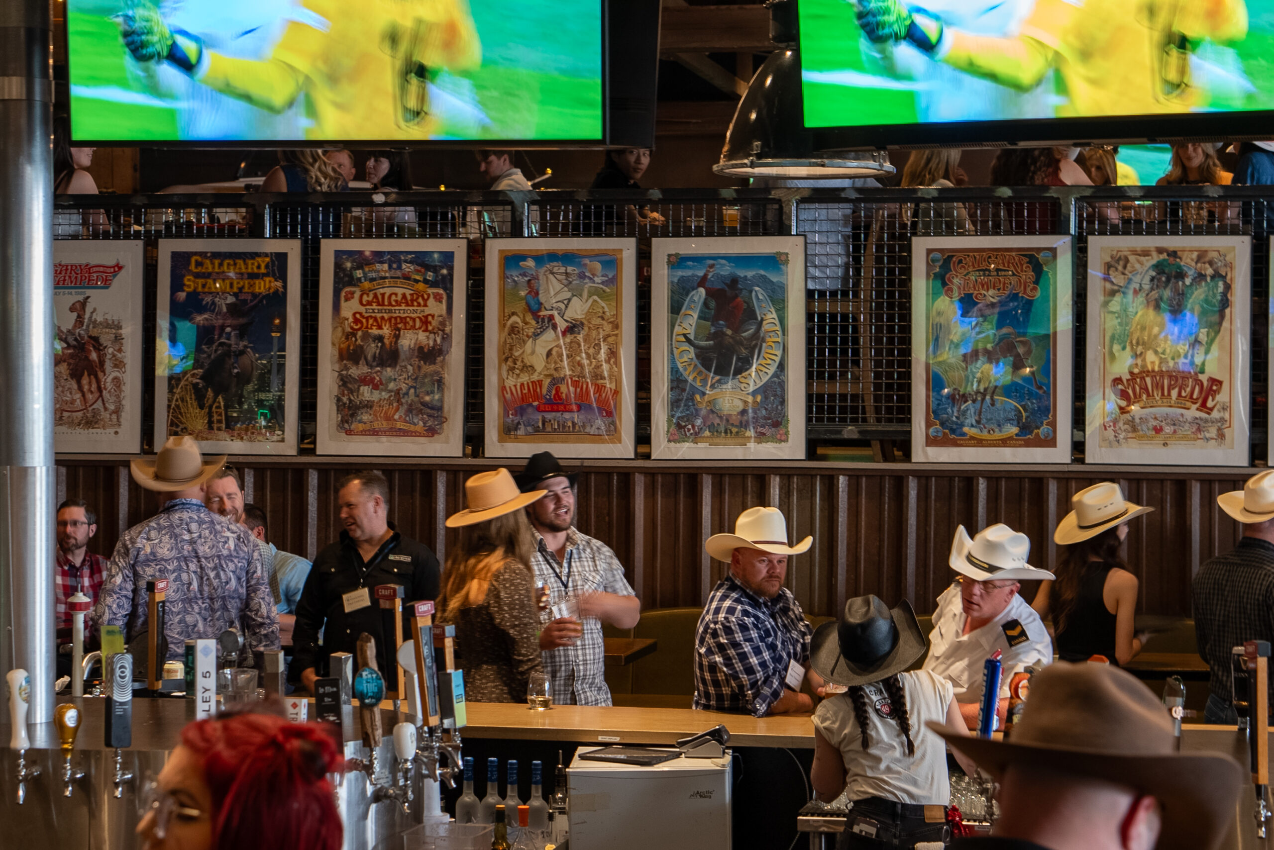 CRAFT Beer Market People in cowboy hats socialize at a bar, with craft beer taps in the foreground and Calgary Stampede posters on the wall. Two large TVs show a sports game above—perfect for group bookings seeking an authentic local vibe.