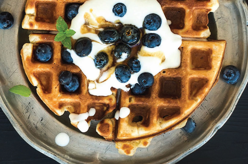 CRAFT Beer Market A plate with quartered waffles topped with blueberries, yogurt, a drizzle of syrup, and a sprig of mint, all made with locally sourced ingredients for the freshest flavor.