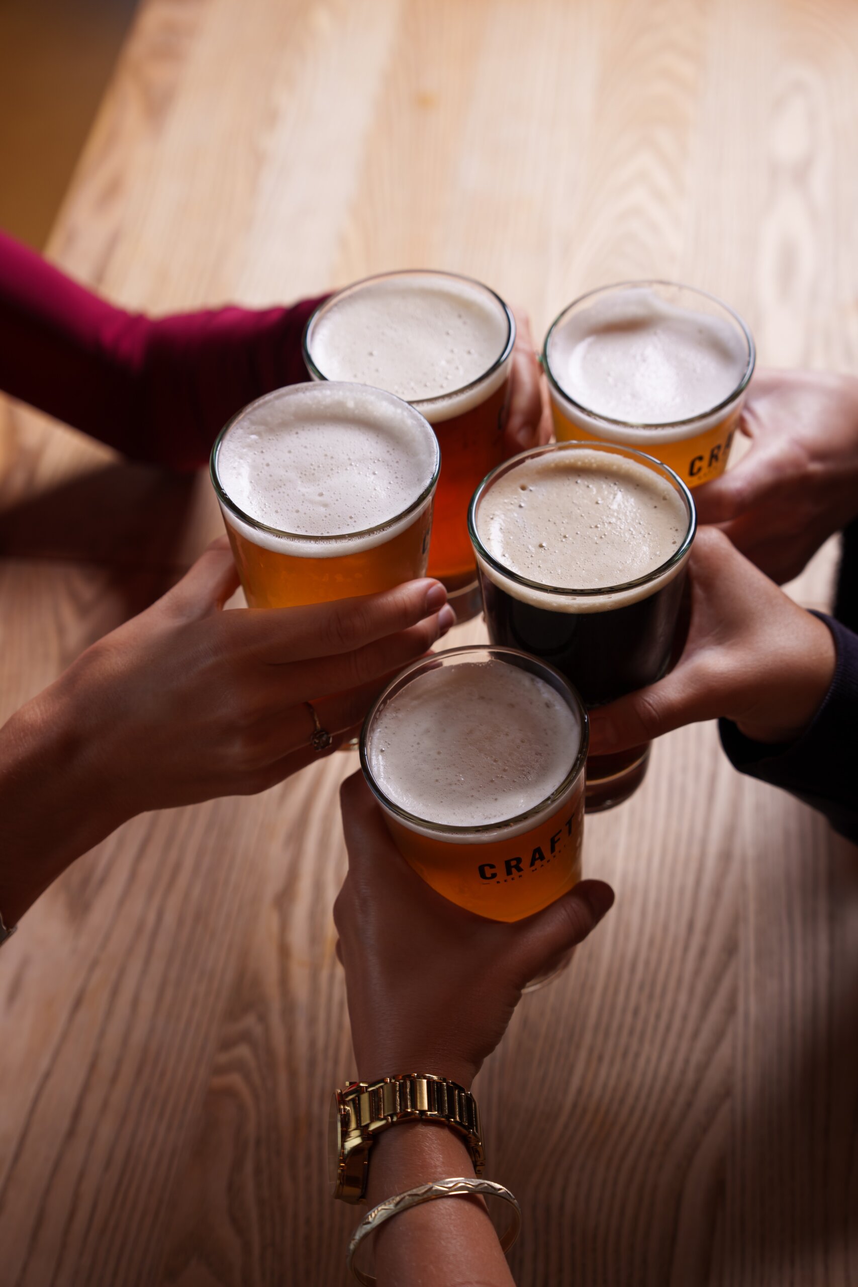 CRAFT Beer Market Five people clinking glasses of craft beer together over a wooden table, celebrating good times with locally sourced ingredients.