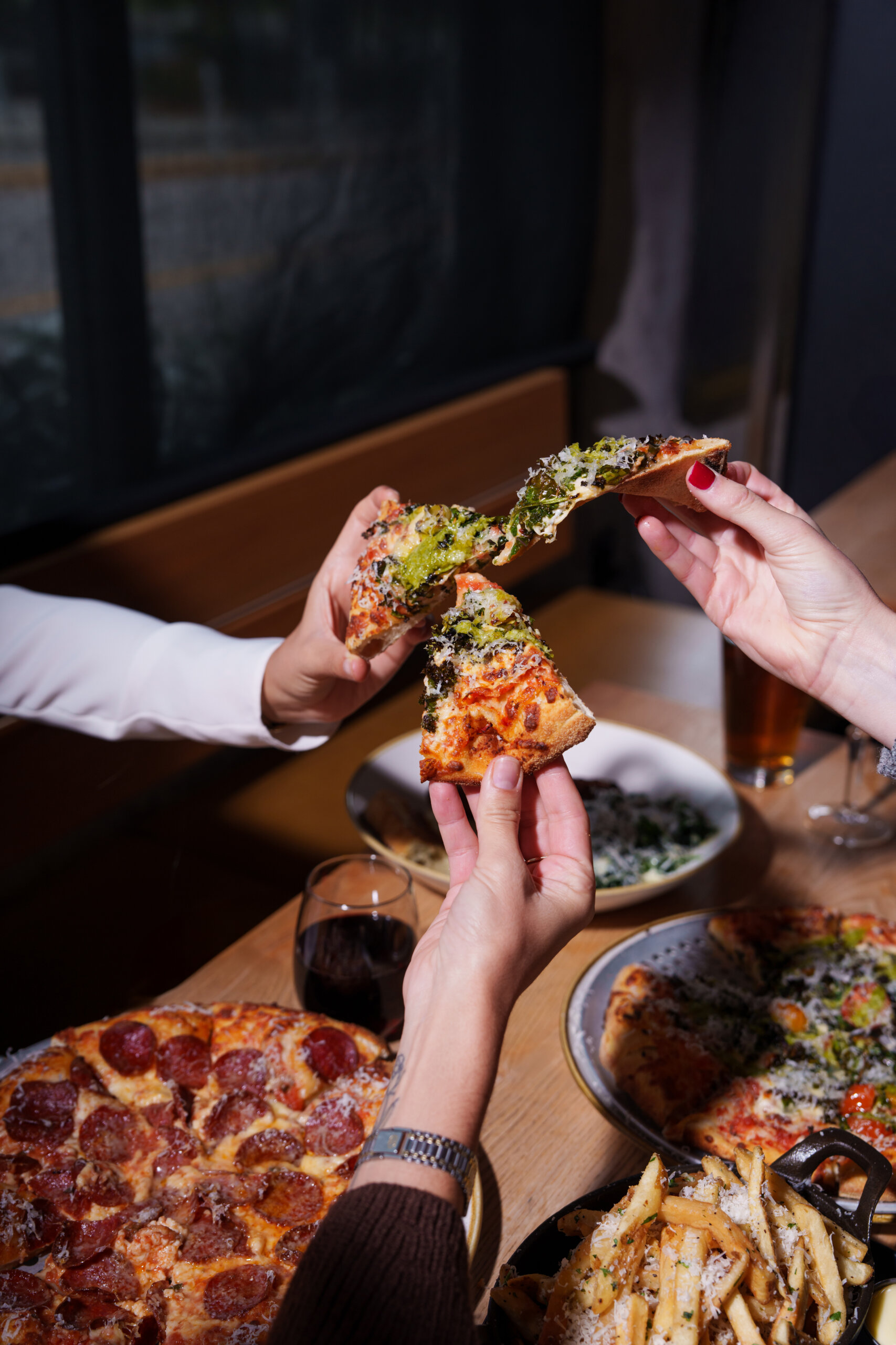 CRAFT Beer Market Three people hold up slices of pizza over a table with more pizza, fries, craft cocktails, a glass of wine, and a drink—sharing food in a lively restaurant setting. Perfect spot for group bookings or private events.