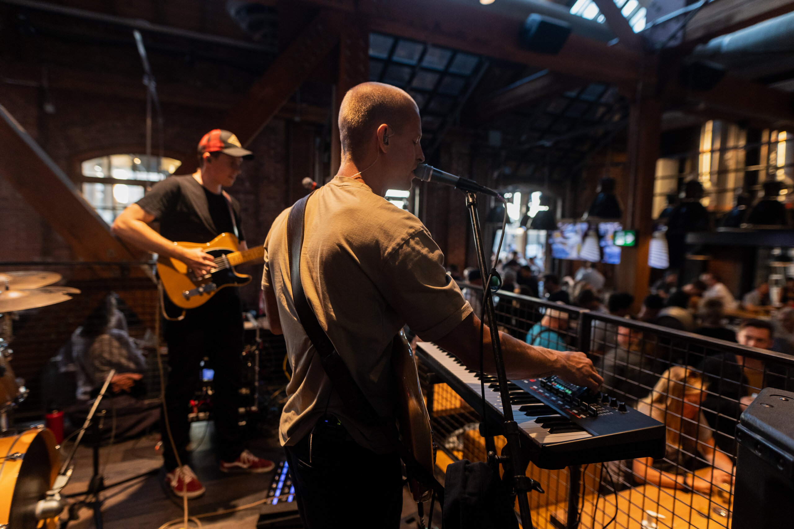 CRAFT Beer Market Two musicians perform indoors; one plays keyboard and sings into a microphone, while the other plays electric guitar. Guests enjoy craft cocktails as the audience sits at tables in the background.
