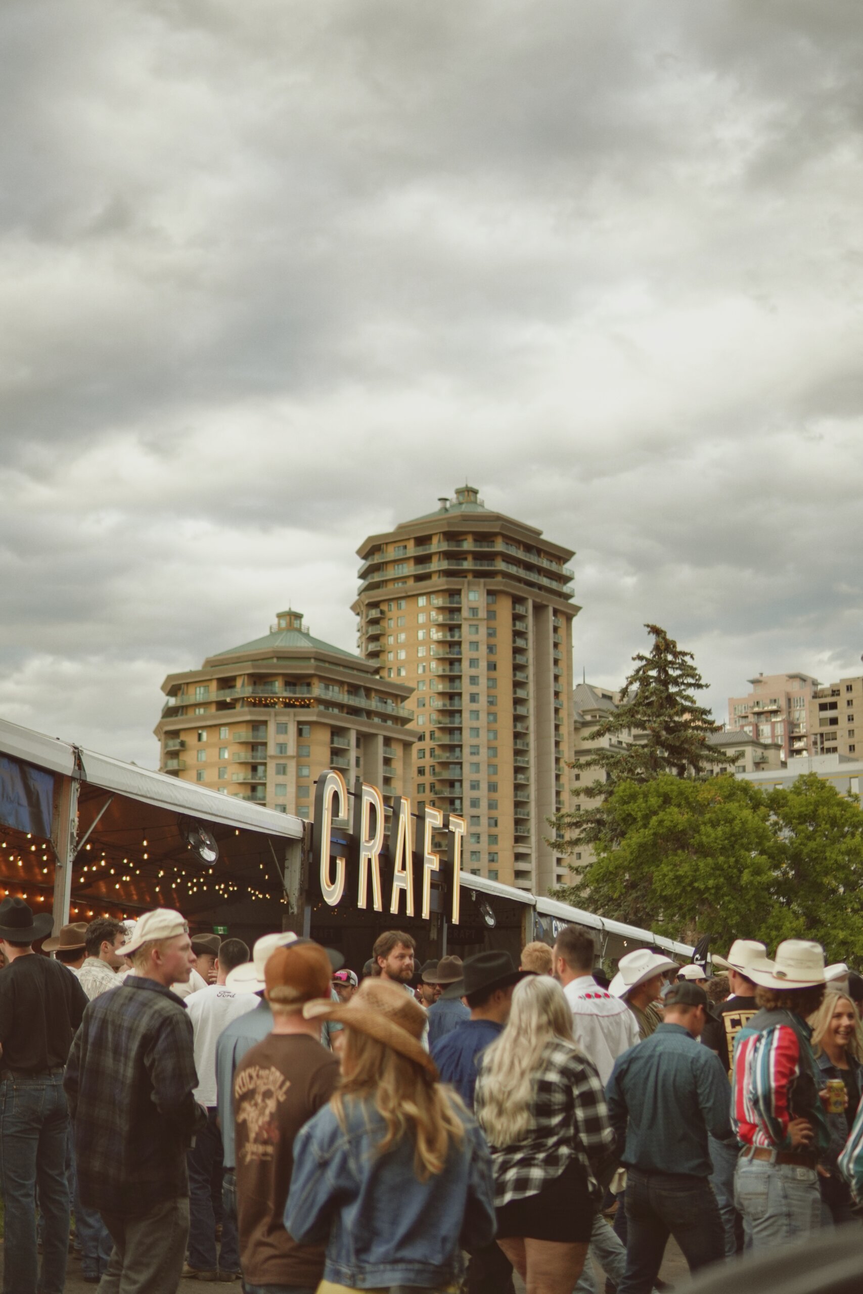 CRAFT Beer Market A crowd gathers outside a tent with a "CRAFT" sign at an outdoor event, where craft cocktails are enjoyed beneath cloudy skies and towering buildings.