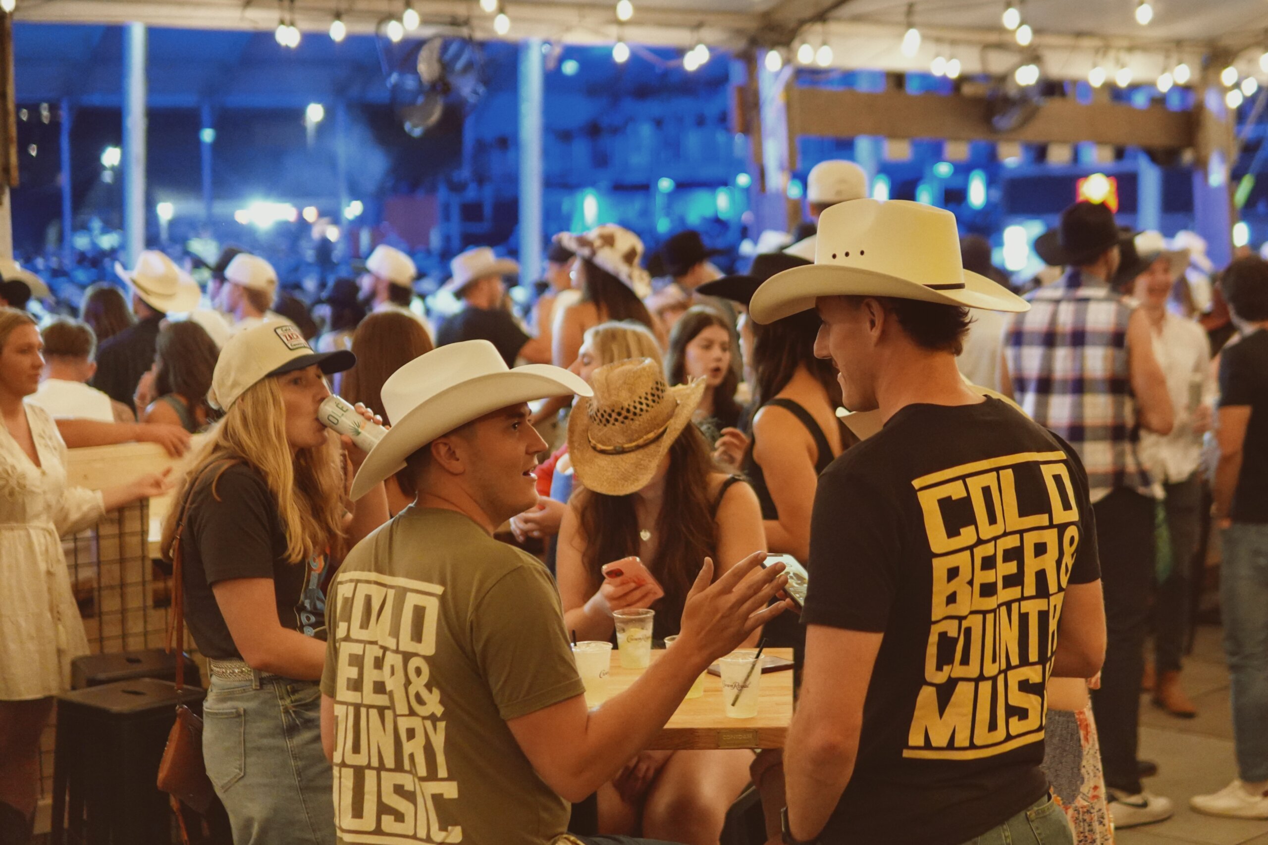 CRAFT Beer Market Crowd of people in cowboy hats and casual clothes gather at an indoor-outdoor venue; two men wear shirts reading "Cold Beer & Country Music" as friends sip craft beer and enjoy the lively atmosphere.