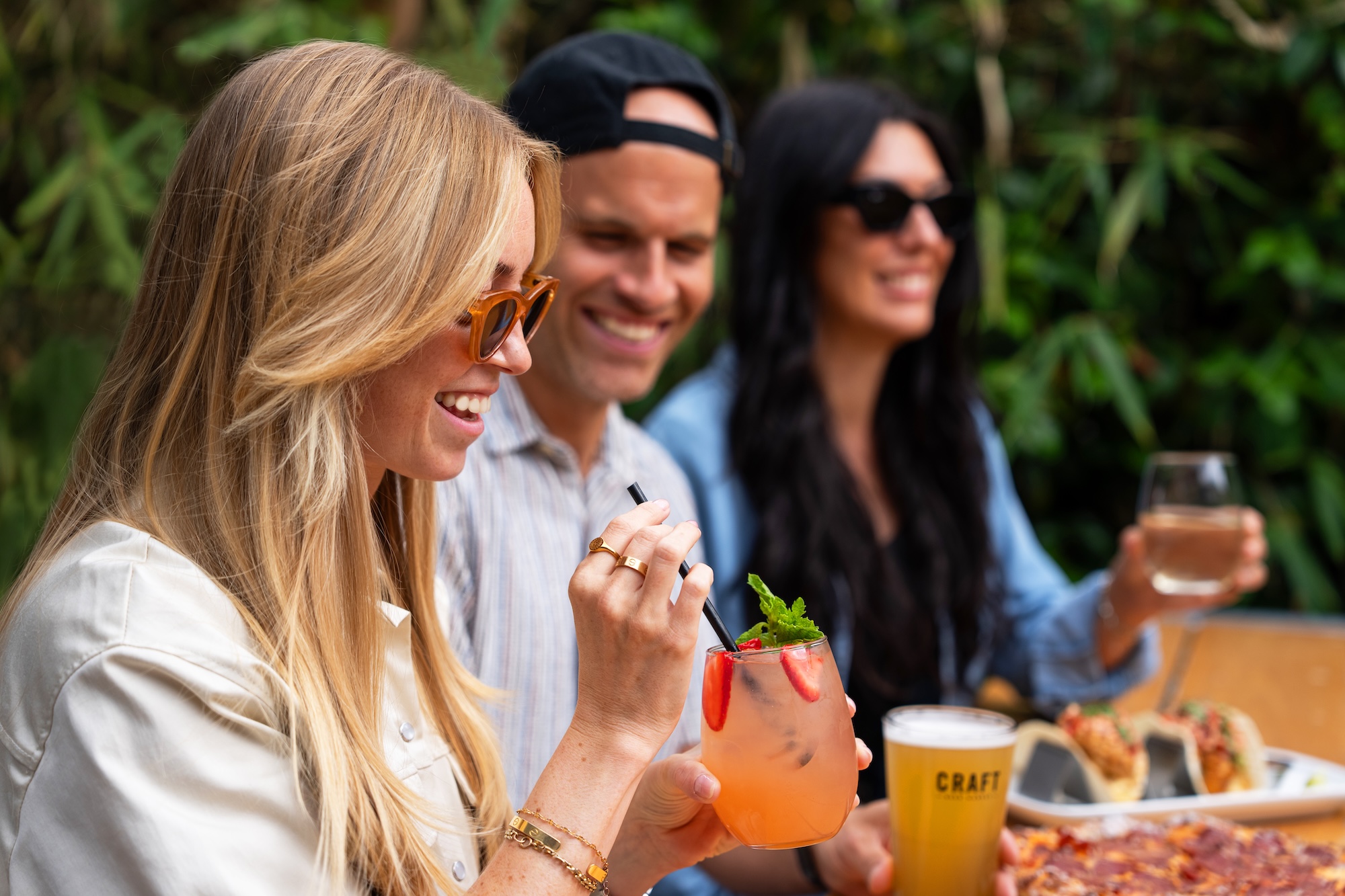 CRAFT Beer Market Three people sit outdoors at a table with drinks and food, smiling and socializing. One person holds a craft cocktail garnished with mint and strawberries, highlighting the use of locally sourced ingredients.