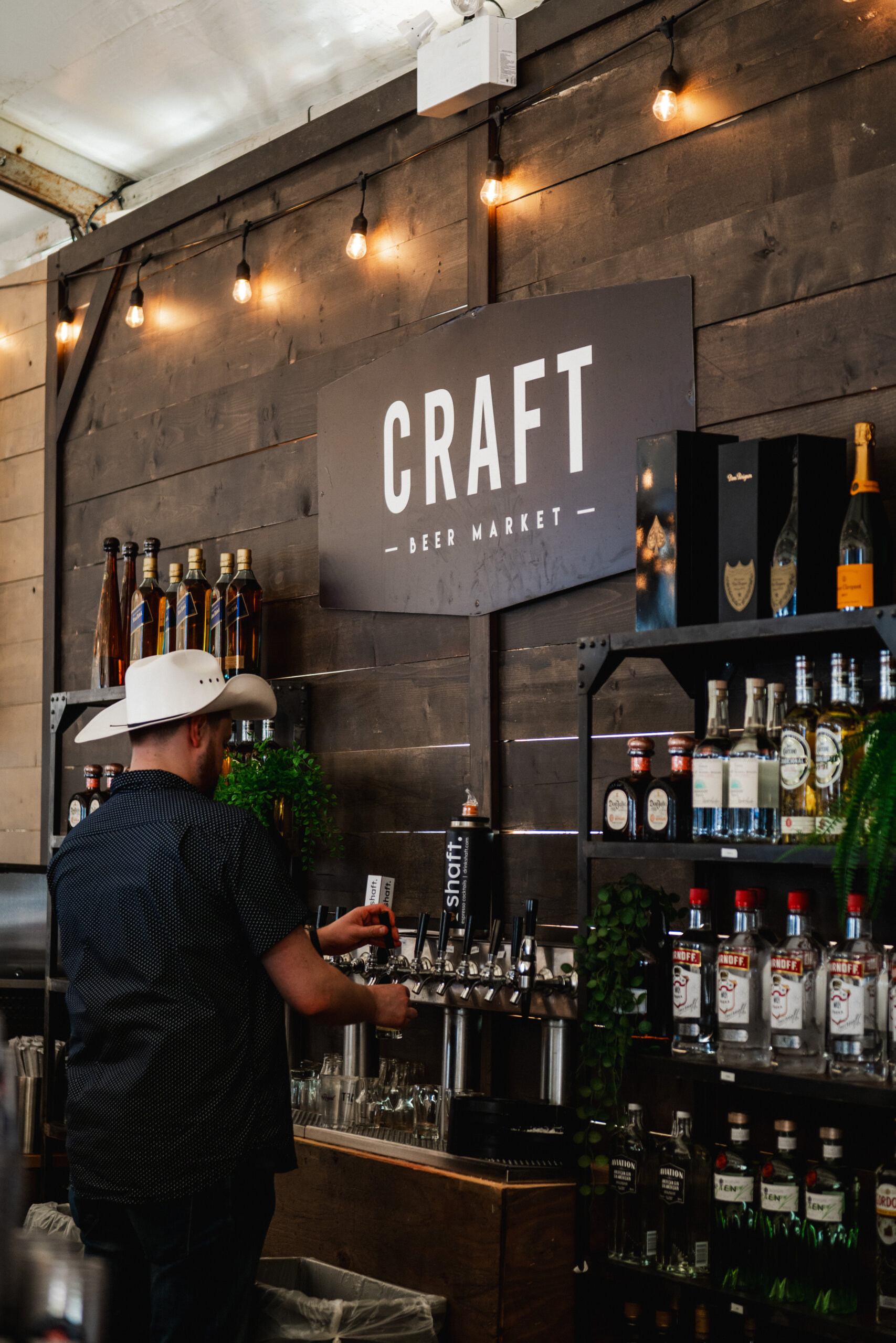 CRAFT Beer Market A person in a cowboy hat pours a drink at a bar with shelves of liquor bottles under a sign reading "Craft Beer Market," highlighting the unique flavor of craft beer and the welcoming space for private events.