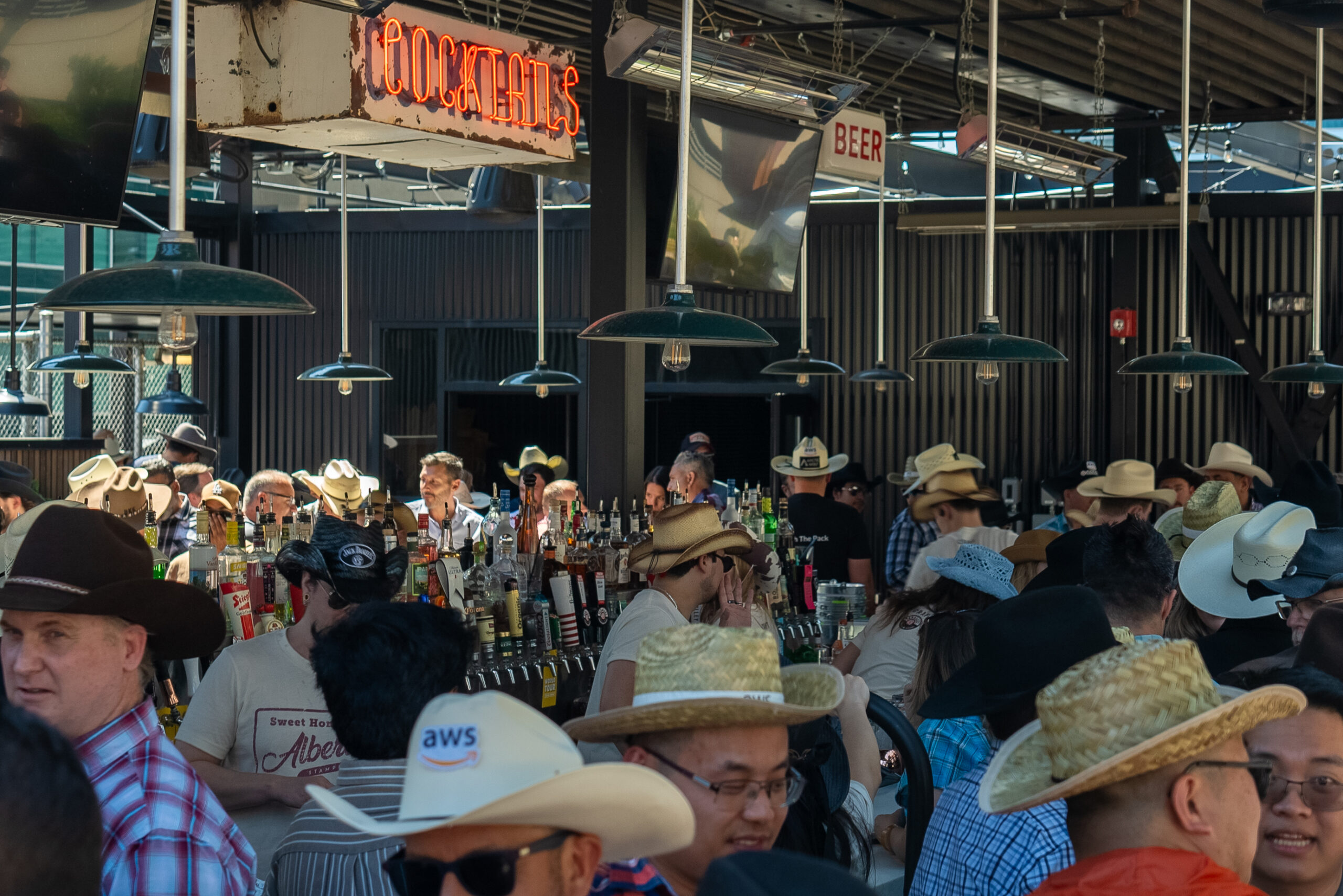 CRAFT Beer Market A crowded bar scene with many people wearing cowboy hats, standing around a bar stocked with bottles of craft beer; signs read “COCKTAILS” and “BEER.”.