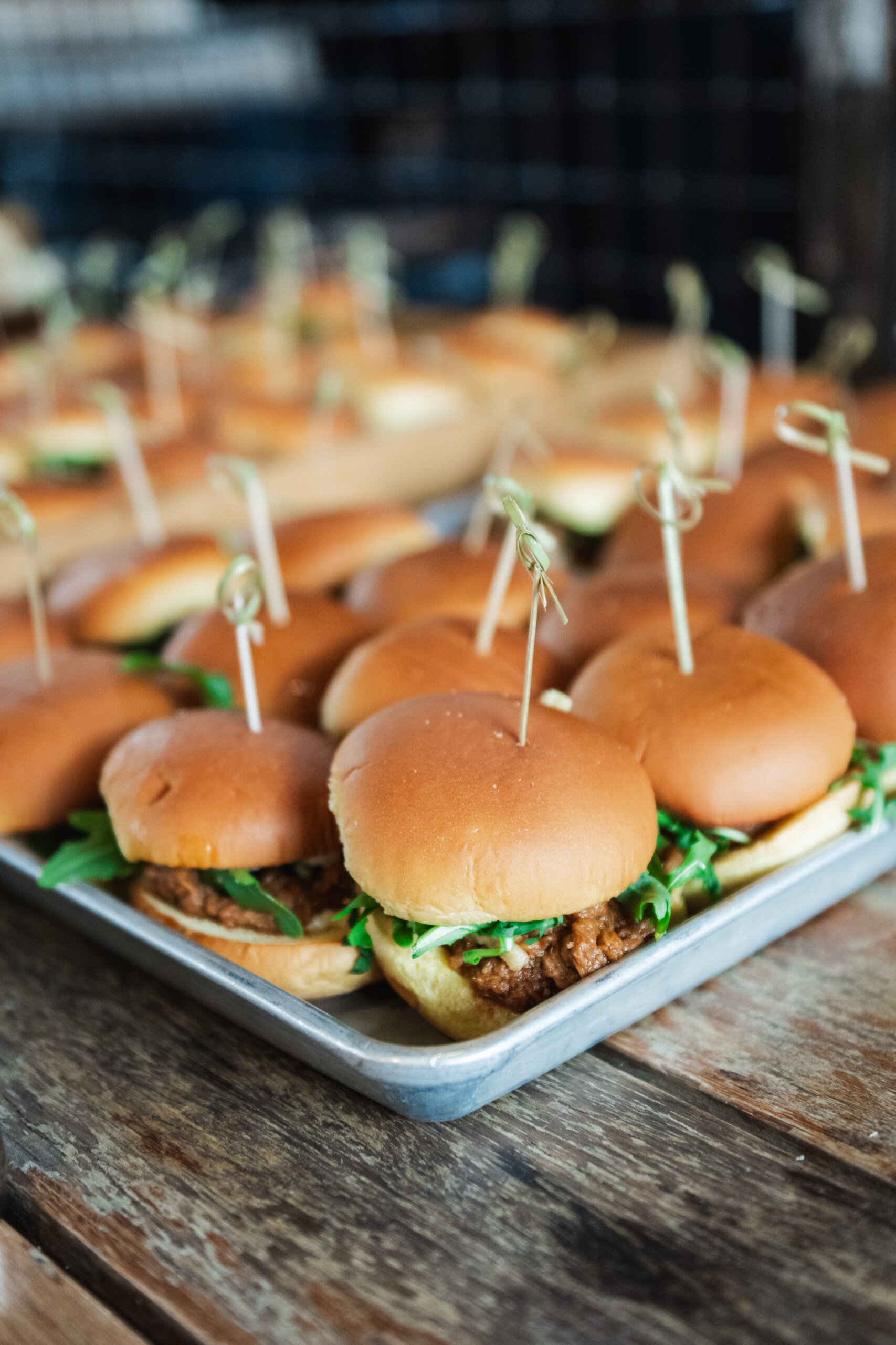 CRAFT Beer Market A tray of small sandwiches with buns, greens, and a filling made from locally sourced ingredients, each held together with a wooden skewer, on a wooden table.