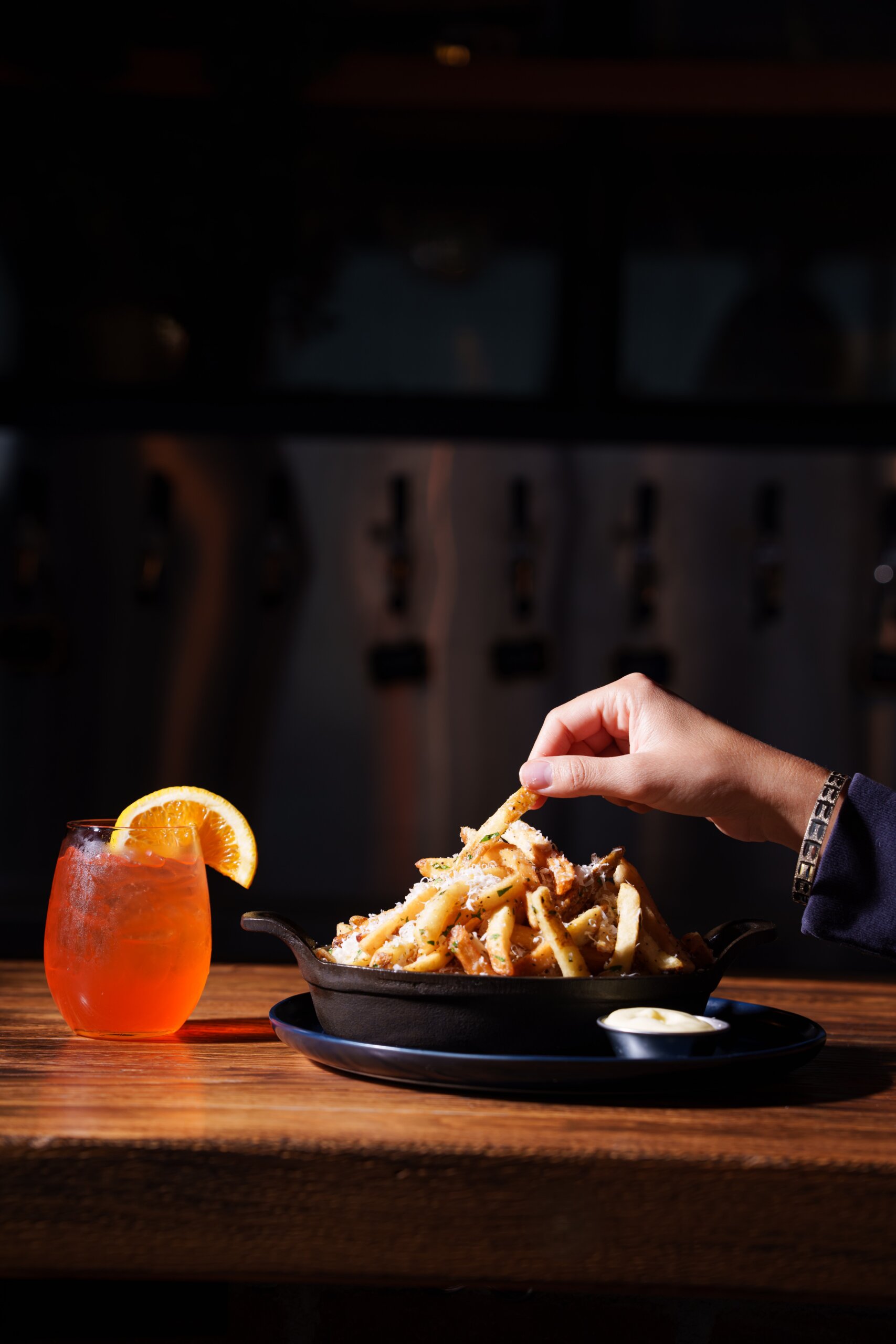 CRAFT Beer Market A hand reaches for loaded fries in a skillet on a wooden table, accompanied by a glass of craft cocktails garnished with orange and a small cup of sauce beside it.