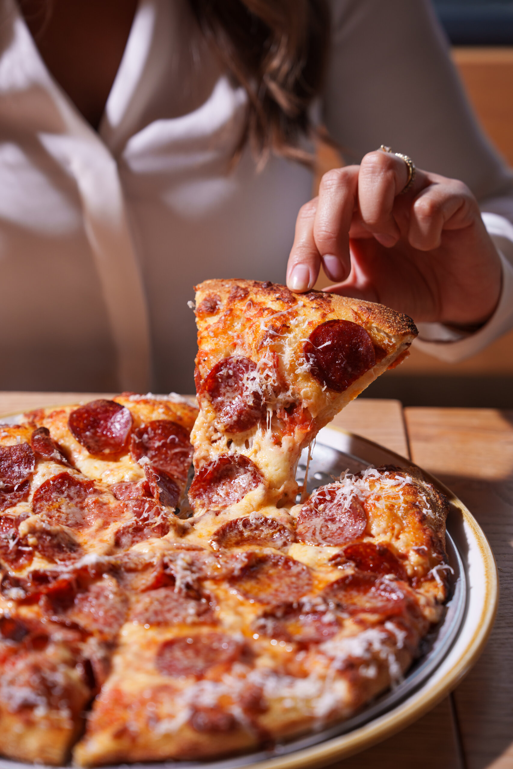 CRAFT Beer Market A person in a white shirt lifts a slice of pepperoni pizza from a whole pizza on a plate, ready to enjoy alongside craft beer and good social vibes.