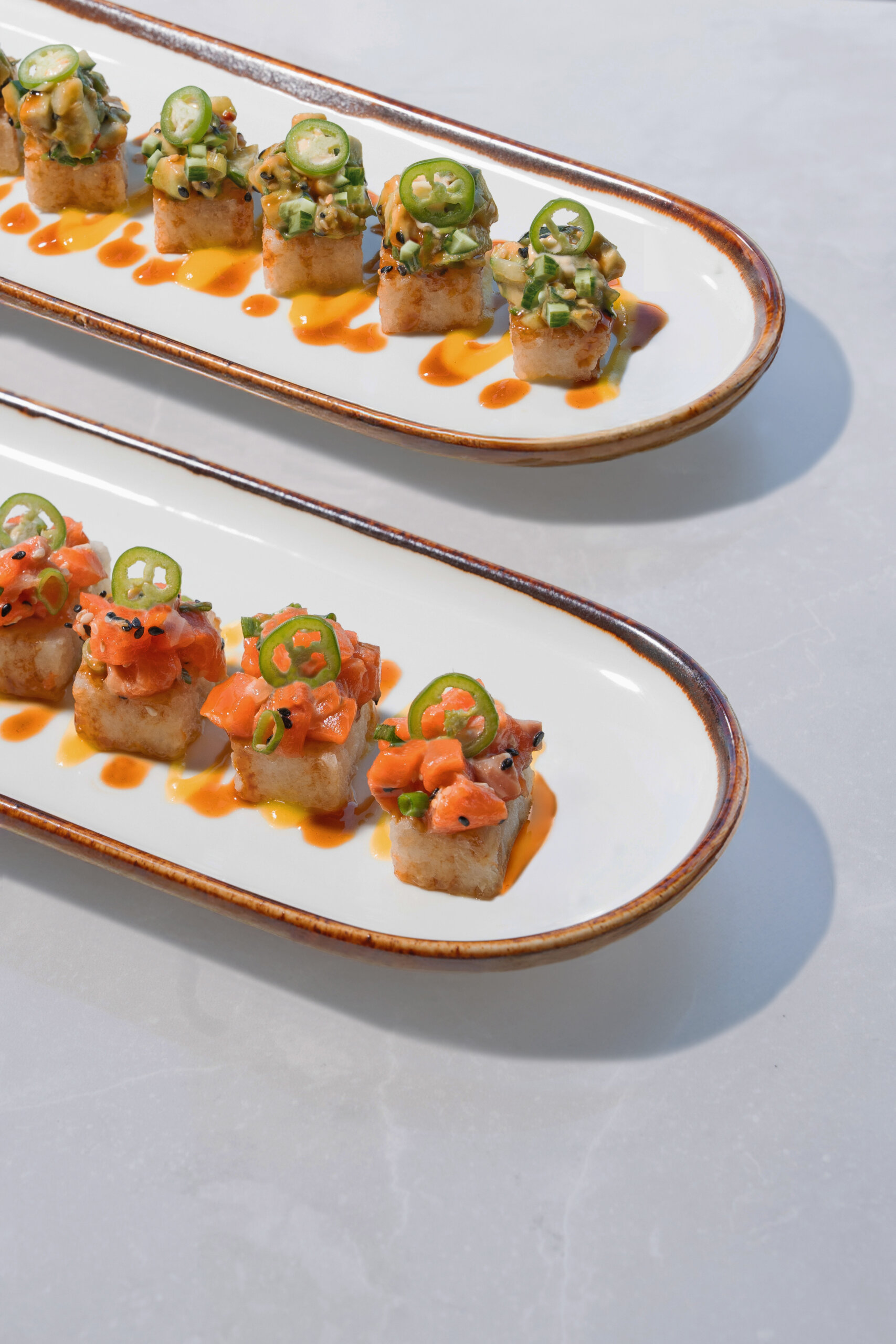 CRAFT Beer Market Two long oval plates each hold four pieces of crispy rice topped with diced fish, sliced jalapeños, and sauce—perfect bites to enjoy with craft cocktails and social vibes on a light surface.