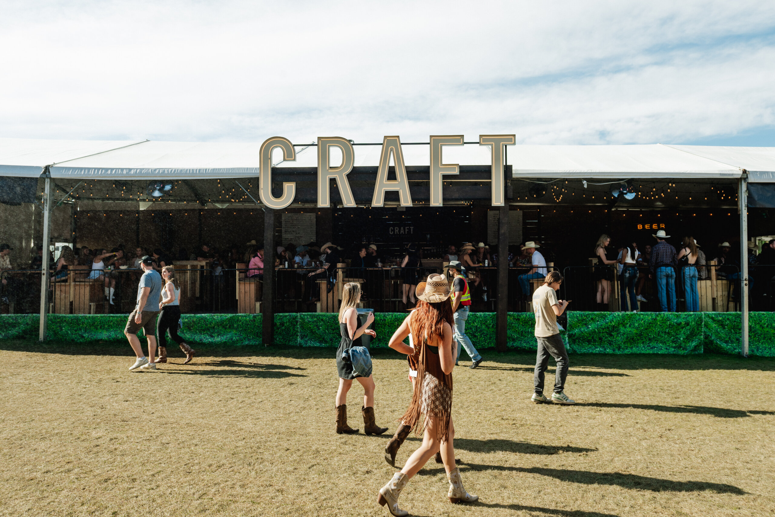 CRAFT Beer Market People walk in front of a large tent labeled CRAFT at the Cowboys Music Festival on a sunny day. Some are dressed in casual festival attire.