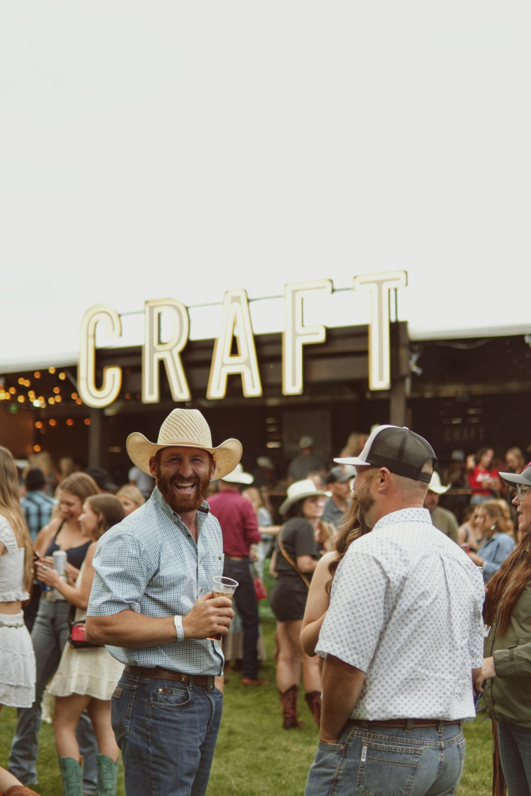 CRAFT Beer Market People in casual western attire gather outdoors in front of a large illuminated CRAFT sign, with one man smiling at the camera while holding a drink—capturing the lively spirit of Cowboys Music Festival.