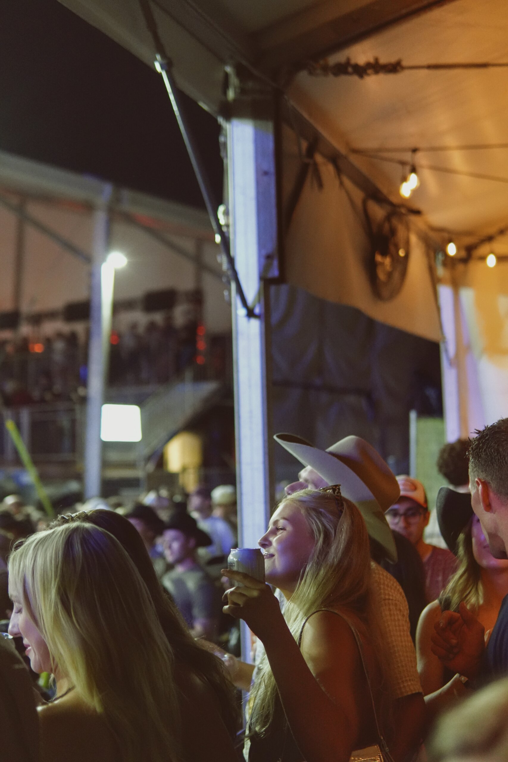 CRAFT Beer Market People gathered at an indoor-outdoor Cowboys Music Festival at night, some wearing hats, one woman in the foreground holding a drink and looking toward the stage.