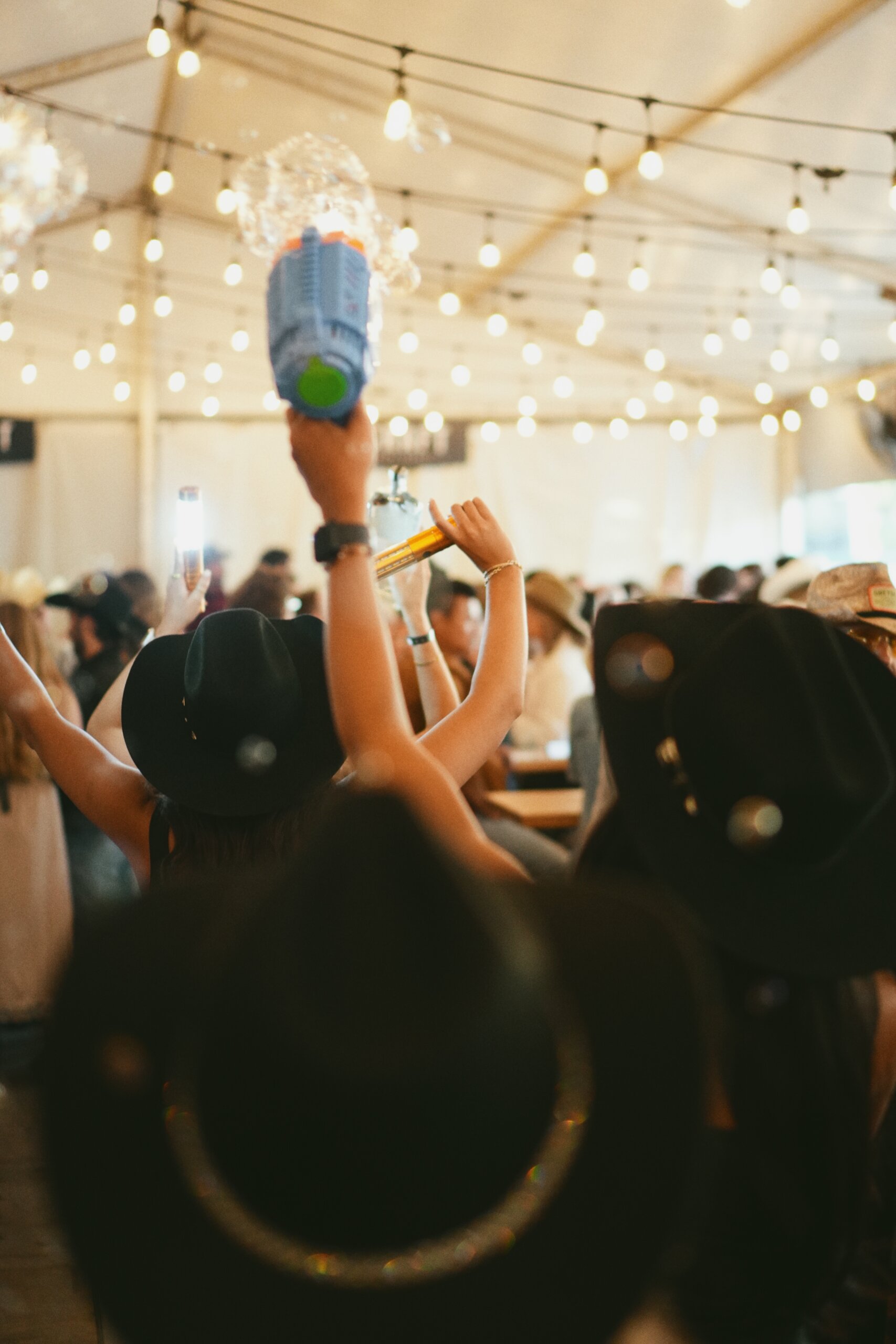 CRAFT Beer Market People wearing black hats gather indoors under string lights at Cowboys Music Festival; one person raises a toy water gun and a drink, facing away from the camera in a festive crowd.