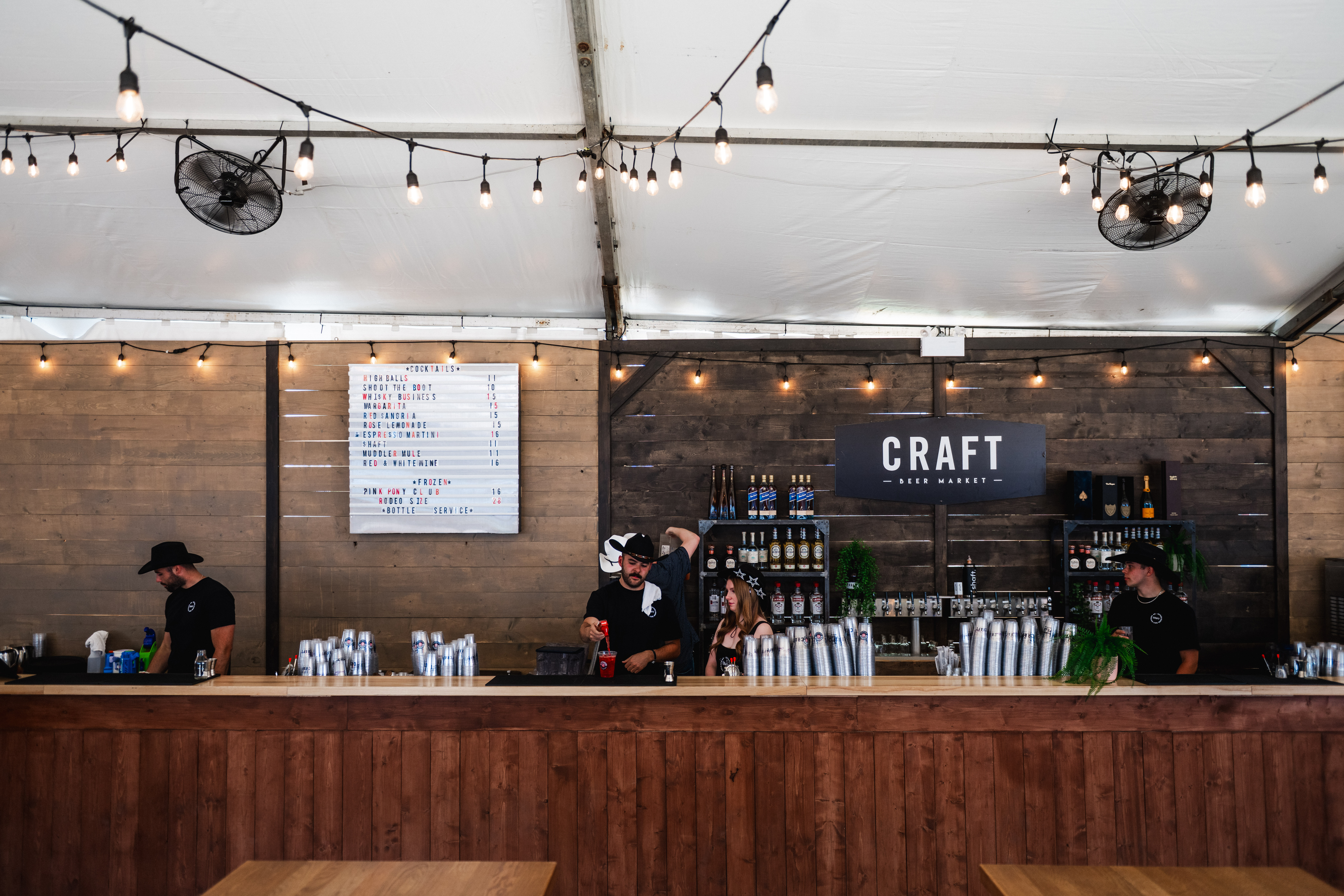 CRAFT Beer Market Bartenders work behind a large wooden bar at CRAFT, under string lights with a Cowboys Music Festival vibe, a drink menu, and shelves of bottles in the background.