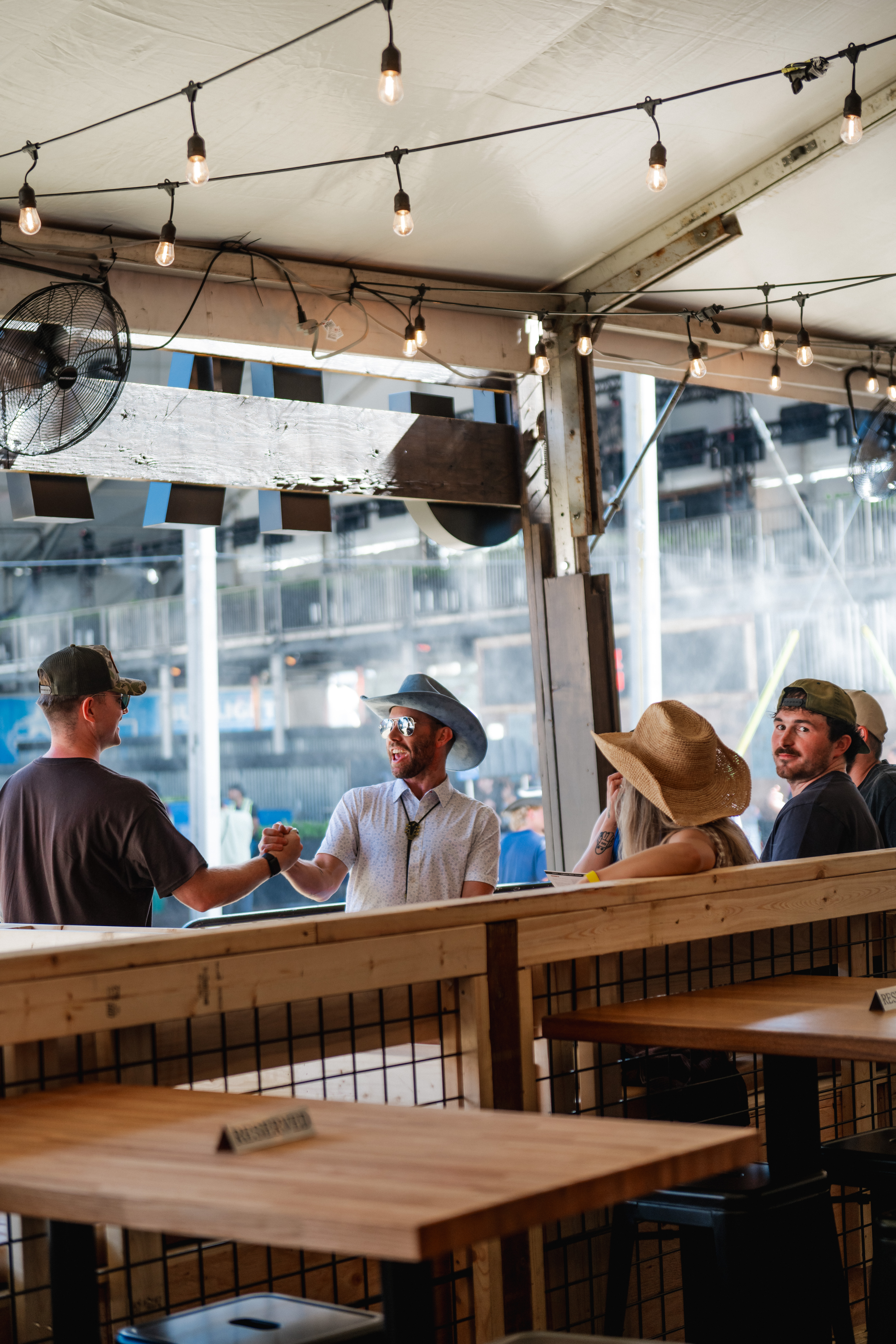 CRAFT Beer Market A group of people wearing hats sit and stand around a wooden table in an outdoor covered area at the Cowboys Music Festival, with string lights overhead.