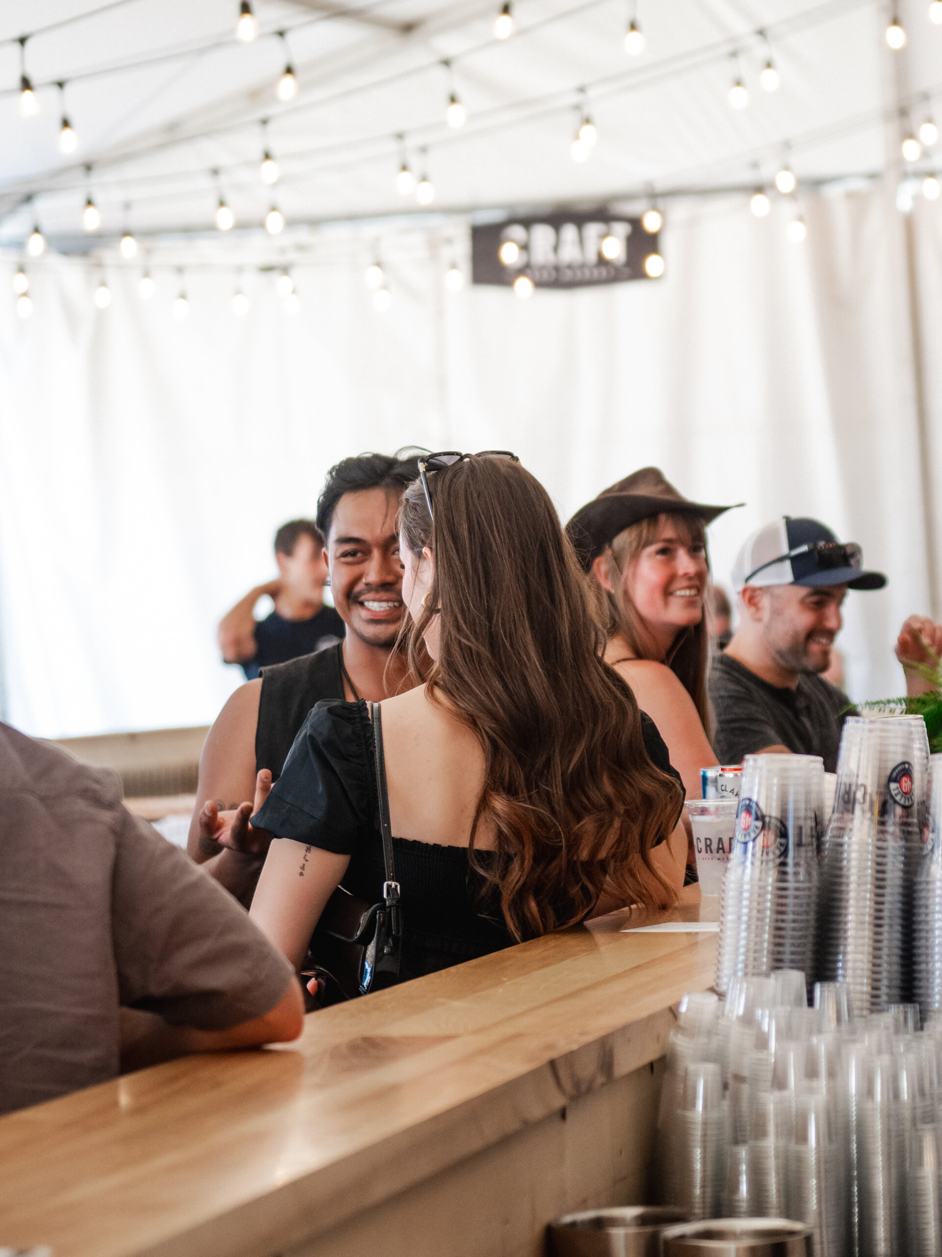 CRAFT Beer Market People converse and smile at a bar counter under string lights at Cowboys Music Festival, with plastic cups stacked in the foreground and a bright CRAFT sign visible in the background.