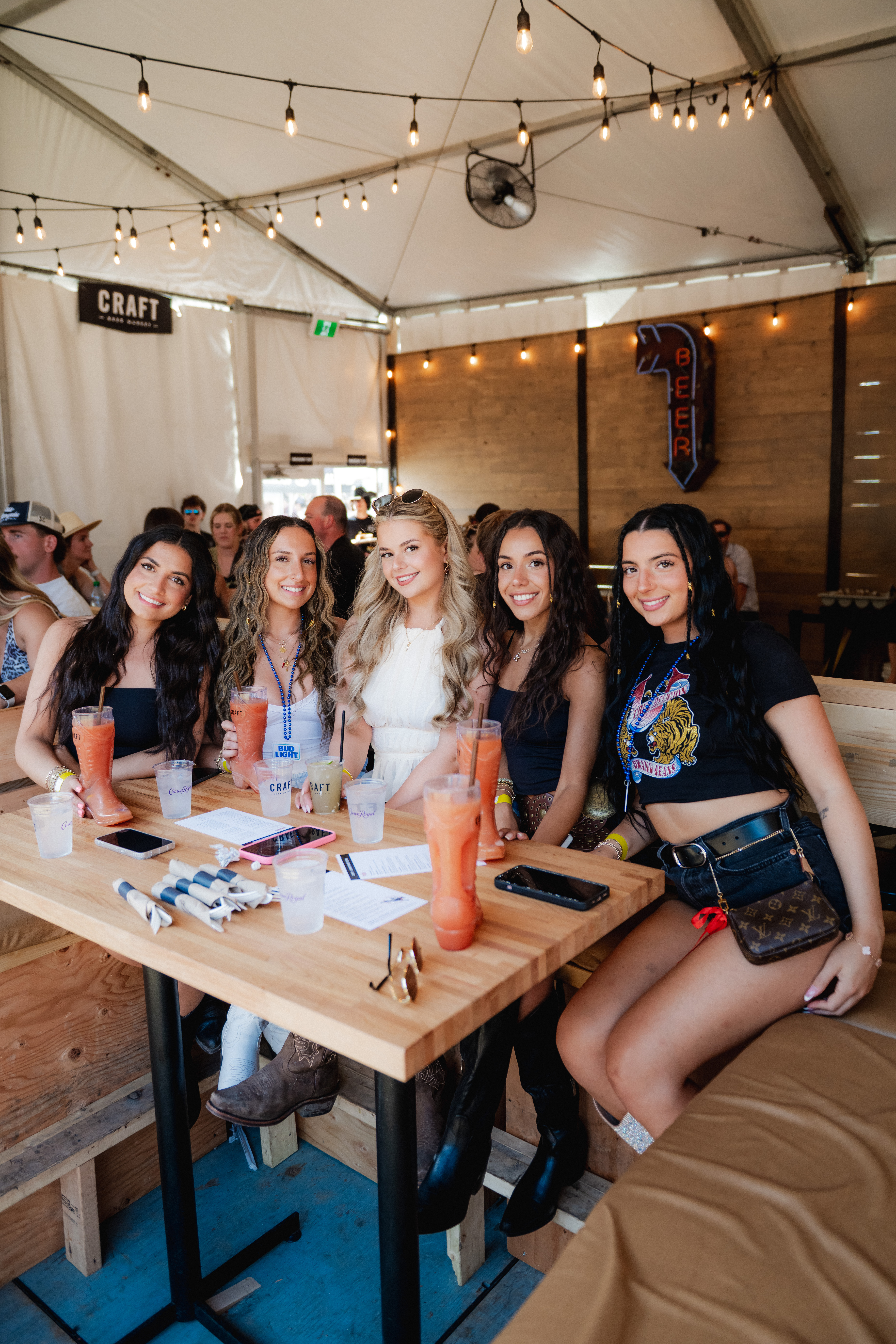 CRAFT Beer Market Five women sit at a wooden table with drinks in a lively indoor venue during the Cowboys Music Festival, smiling at the camera; people and string lights are visible in the background.