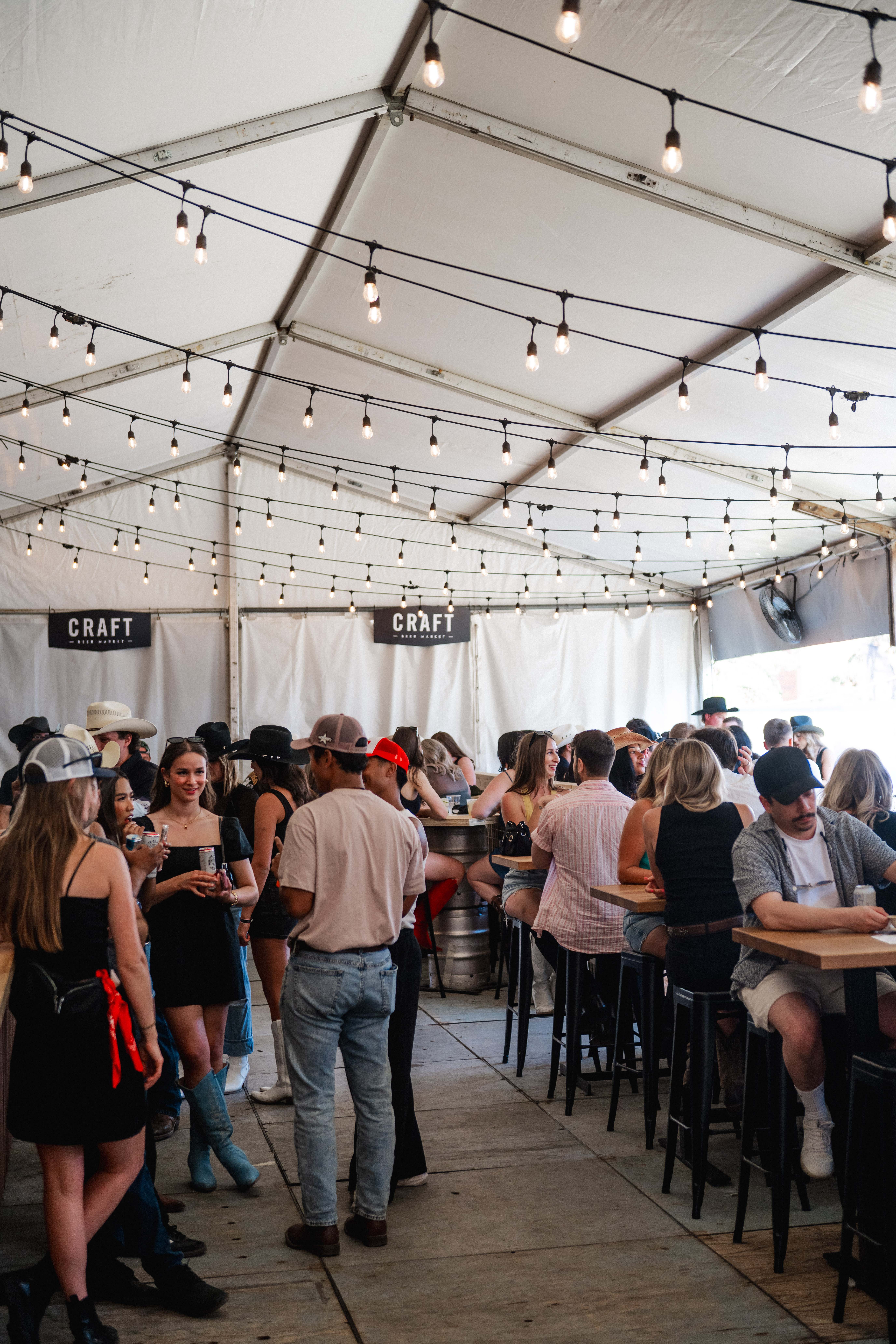 CRAFT Beer Market People gather and socialize under a white tent decorated with string lights at the lively Cowboys Music Festival bar area labeled CRAFT. Some guests stand while others are seated at high tables, enjoying the vibrant music festival atmosphere.