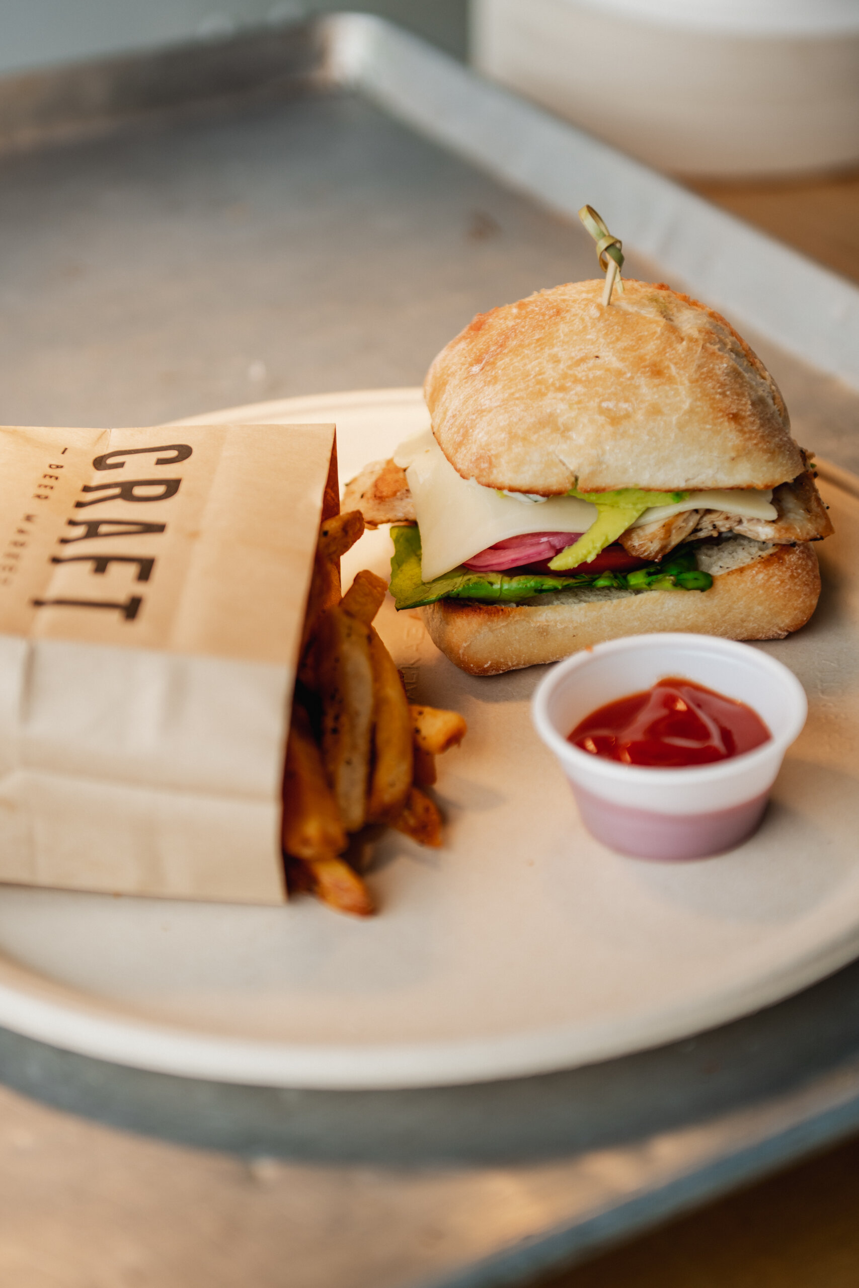 CRAFT Beer Market A CRAFT sandwich with cheese, lettuce, tomato, and chicken on a bun, served with fries in a paper bag and a small cup of ketchup on a white plate—perfect for fueling up before the Cowboys Music Festival.